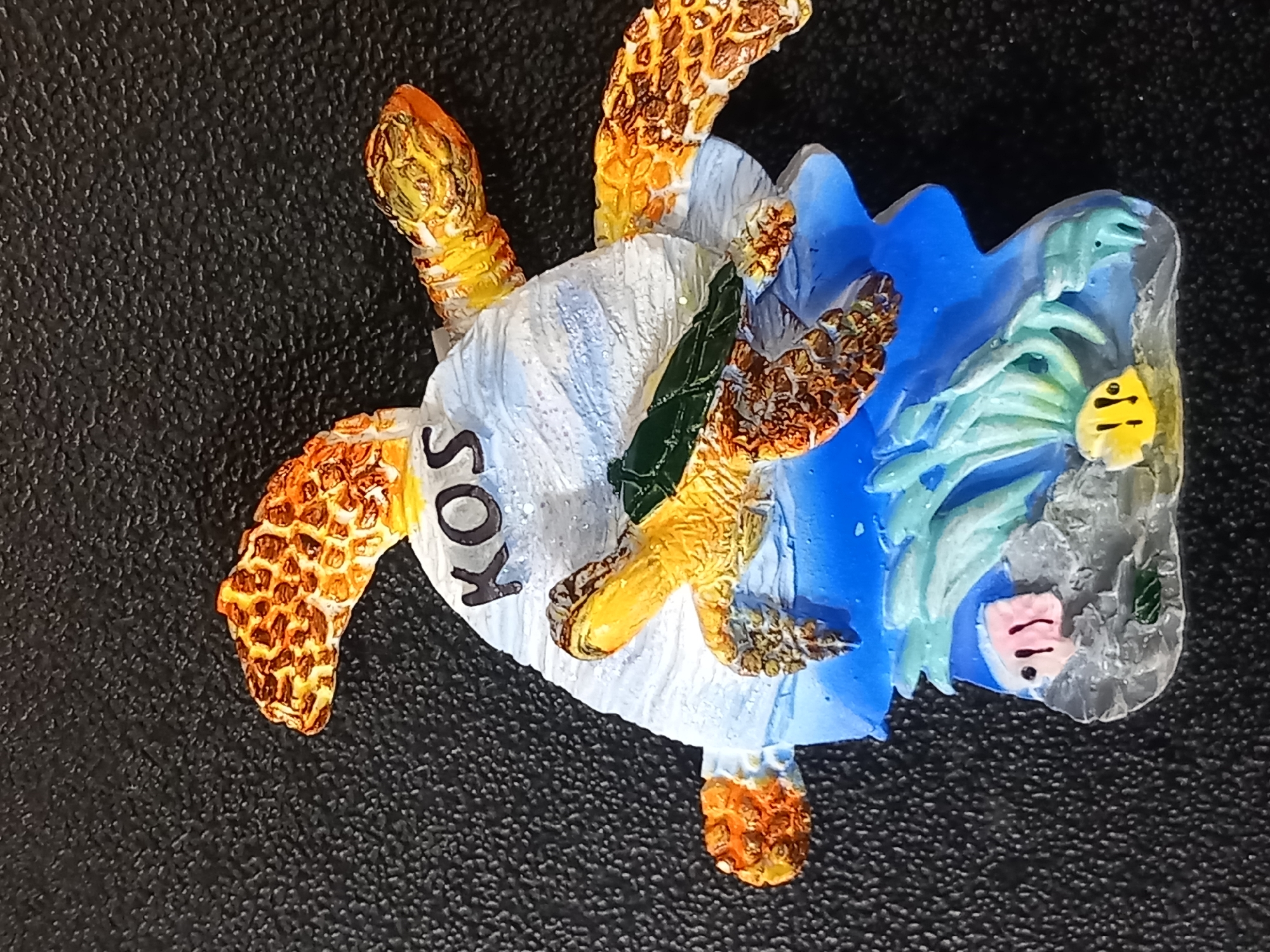 A souvenir magnet depicts a detailed, three-dimensional scene of sea turtles and underwater flora. The magnet features a stylized representation of a sea turtle with its shell adorned with the letters "KOS" in black. The turtle is positioned atop a blue, wave-like structure, which appears to be a base for the magnet. The base is further enhanced with colorful representations of aquatic plants and two small fish. One fish is pink with black dots for eyes, and the other is yellow with black dots. The background of the image is a textured black surface, possibly a countertop or a fabric. The lighting is direct, casting subtle shadows on the magnet's surface.