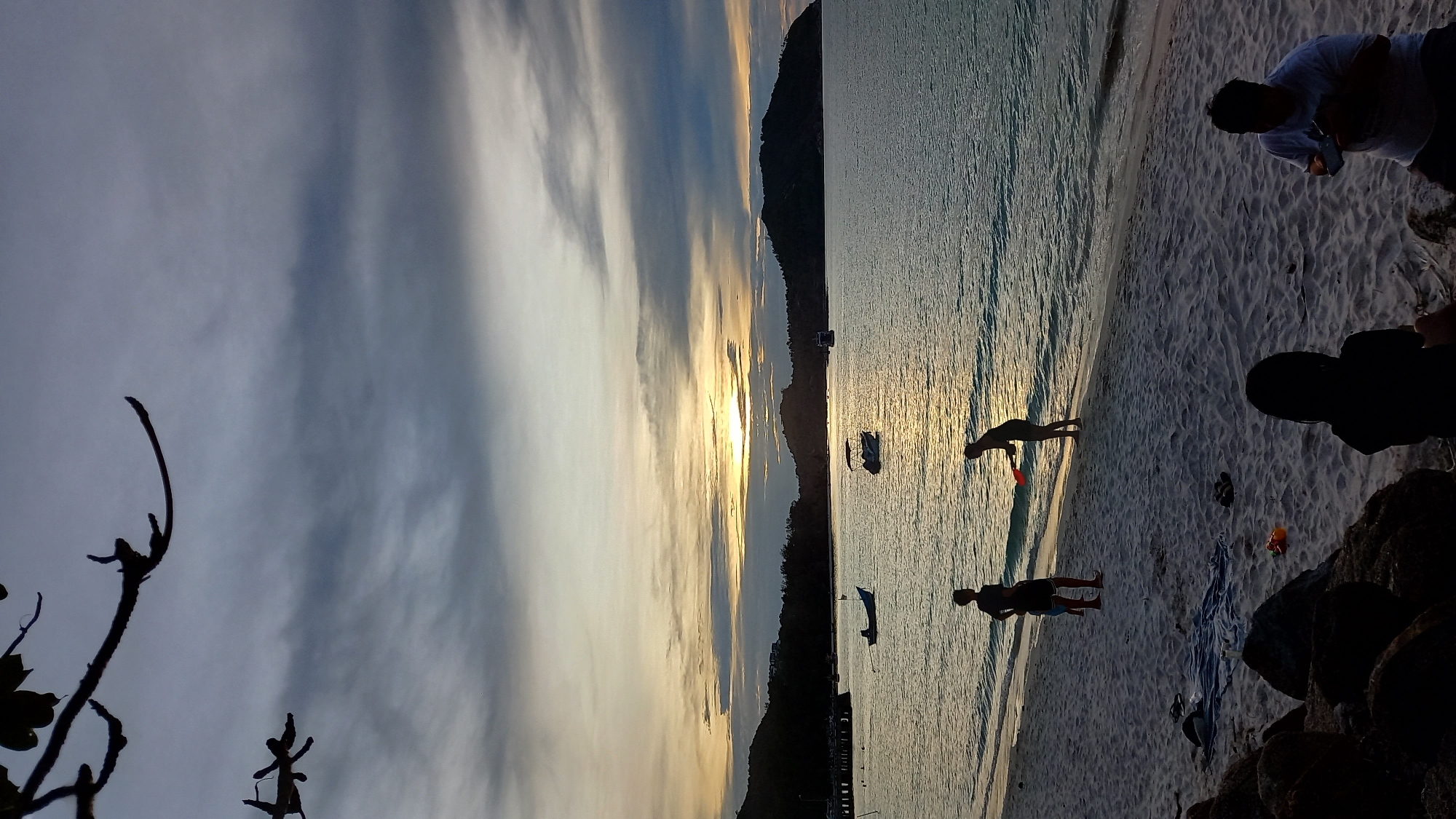 The image captures a beach scene at sunset in ตำบลเกาะพะงัน, Thailand. Silhouettes of people are visible on the sandy shore. In the foreground, two figures are seated on rocks, one appearing to look at a phone, and another facing away from the camera. Further up the beach, two figures stand near the water's edge, one holding a red object. The ocean stretches out to the horizon, with several boats anchored offshore. Islands with lush vegetation form the backdrop, silhouetted against a sky filled with dramatic clouds and the warm glow of the setting sun. Tree branches in the upper left corner frame the top of the image.