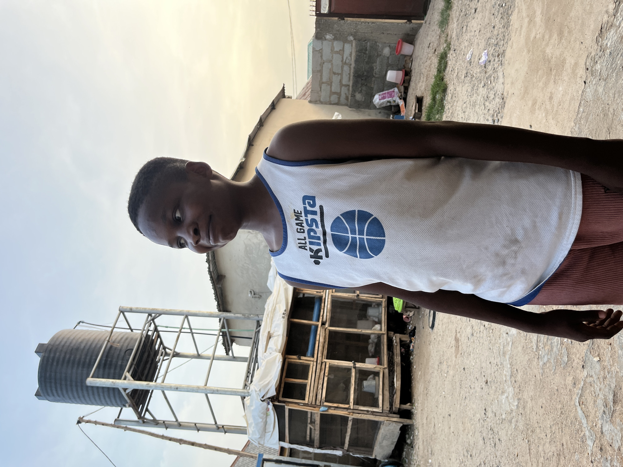 A young man stands outdoors, likely in the city of Iddah, Nigeria. He is wearing a white basketball jersey with a blue trim and a graphic of a basketball. His lower body is partially visible, showing reddish-brown pants. Behind him, there's a water tank mounted on a metal scaffolding structure. A wooden structure, possibly a chicken coop or cage, is also visible, partially covered with a white tarp or fabric. In the background, there are buildings made of concrete blocks. The ground is dirt. The sky is a pale blue.