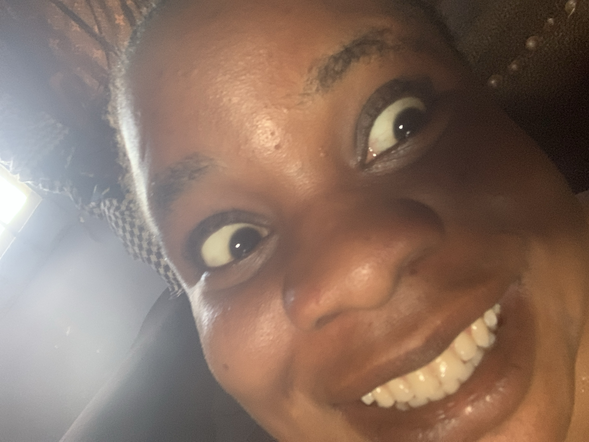 A close-up selfie shows a dark-skinned person smiling broadly, revealing their teeth. Their eyes are wide and slightly upturned. The background is partially obscured, but a checkered pattern can be seen in the upper left corner, and a dark surface with rounded protrusions appears in the upper right corner. The location is unknown.