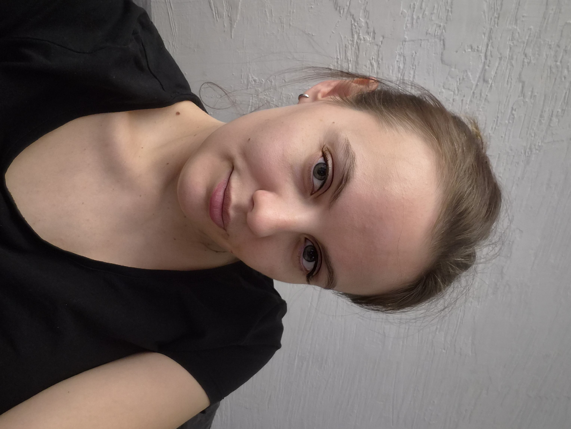 A close-up portrait shows a young woman looking directly at the camera with a slight, gentle smile. Her head is tilted, giving the image a dynamic angle. She has fair skin, striking green or grey eyes, and light brown hair that appears to be pulled back, with a few wispy strands around her face. A small silver earring is visible in her left ear. She is wearing a simple black t-shirt. On her visible collarbone, a small mole is present, along with a few tiny freckles. The background consists of a textured, light grey wall, suggesting an indoor setting. There is no visible text in the image. The overall impression is one of casualness and calm, likely captured in Chaikovsky, Russia.