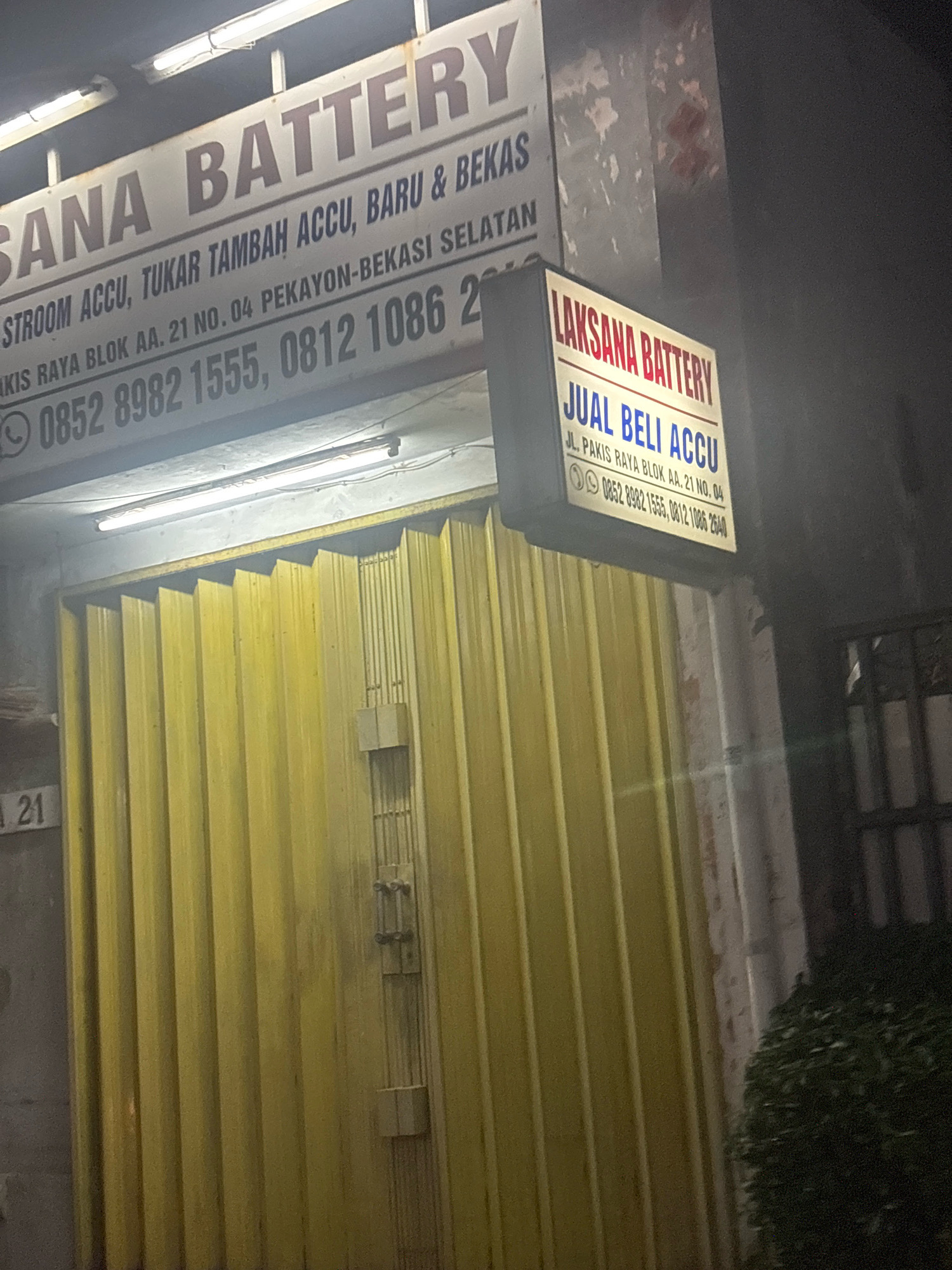 The image shows a nighttime view of a shopfront in Bekasi, Indonesia, identified by the text on two signs. The primary sign, a large horizontal one, reads "SANA BATTERY". Below this, smaller text states, "STROOM ACCU, TUKAR TAMBAH ACCU, BARU & BEKAS" followed by "AKIS RAYA BLOK AA. 21 NO. 04 PEKAYON-BEKASI SELATAN" and a phone number "0852 8982 1555, 0812 1086 2".
A second, smaller, illuminated sign to the right reads "LAKSANA BATTERY" with the text "JUAL BELI ACCU" below it. This sign also displays an address "JL. PAKIS RAYA BLOK AA. 21 NO. 04" and a phone number "0852 8982 1555, 0812 1086 2640". A WhatsApp icon is visible next to the phone numbers on both signs.
The shopfront features a closed, bright yellow, corrugated metal gate. To the left of the gate, a grey concrete wall displays the number "21" in white text. The environment appears to be a street or commercial area with overhead lighting illuminating the signs and shopfront. A dark, dense bush is visible on the far right of the frame. The overall lighting suggests it is dark outside.