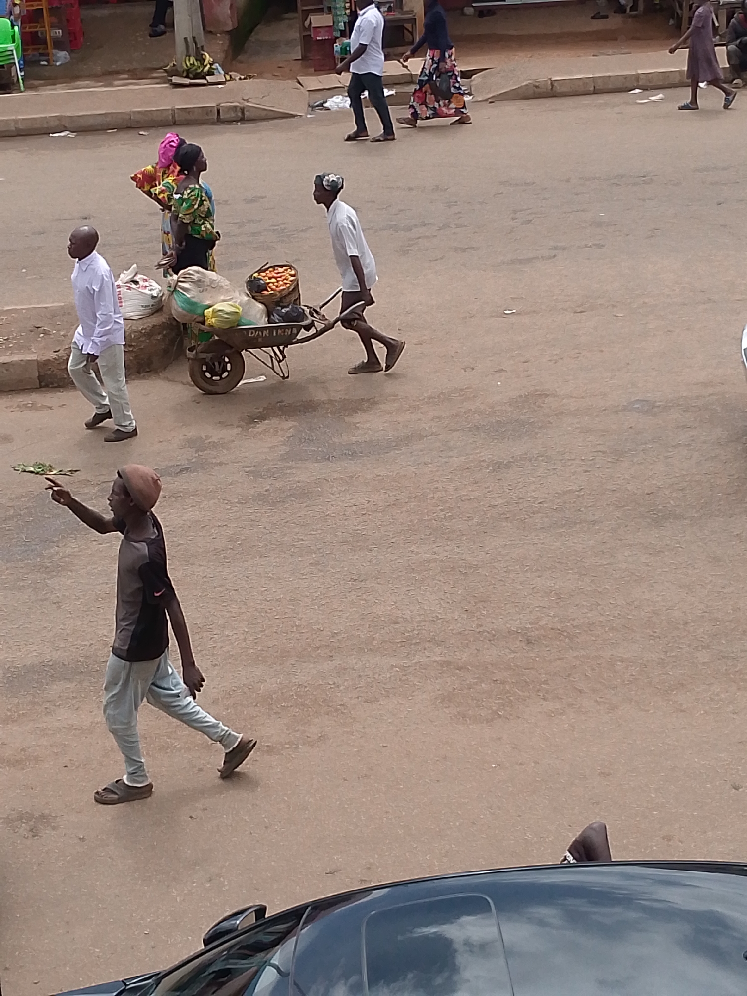 In an outdoor scene set on a dusty, paved street in Bukuru, Nigeria, a man in a dark t-shirt and light-colored pants walks away from the camera, his left arm extended, pointing. He wears a brown beret and brown sandals. To his left, another man in a white shirt and light pants walks towards the camera, his hands clasped behind his back. Behind him, a person in a colorful dress and a pink headscarf stands looking to the right. Beside them, a wheelbarrow is laden with produce, including a basket of red fruits and several bags, one of which is white and has visible lettering. Another person in a white shirt and dark pants pushes the wheelbarrow, walking away from the camera. In the background, a person in a purple dress walks away from the camera. The scene appears to be a busy marketplace or street with several individuals engaged in daily activities.