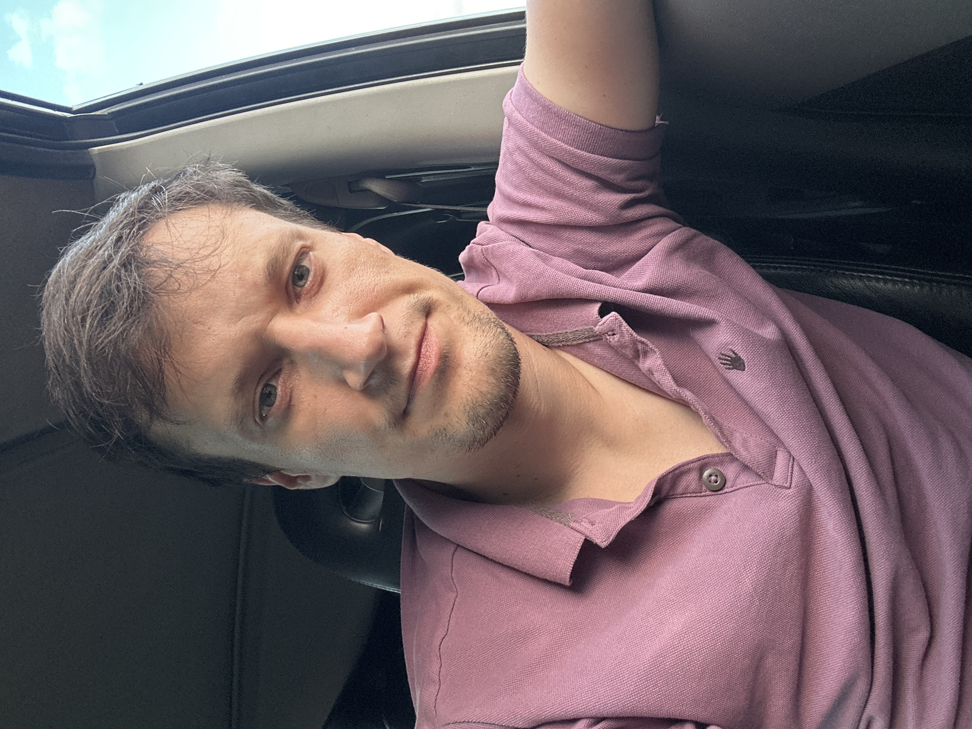 The image is a close-up selfie of a man sitting in a car. He is wearing a pink polo shirt and has short, dark hair. The background shows the interior of the car, including a black leather seat and a portion of the car door. Outside the car, the sky is visible with some clouds. The man is looking directly at the camera with a neutral expression. The lighting suggests it is daytime. The location is not specified within the image itself, but if we assume it is in Makiv, Ukraine, this detail would be external context. The image shows no signs of any specific events or interactions, depicting a still moment of the individual in the car.
