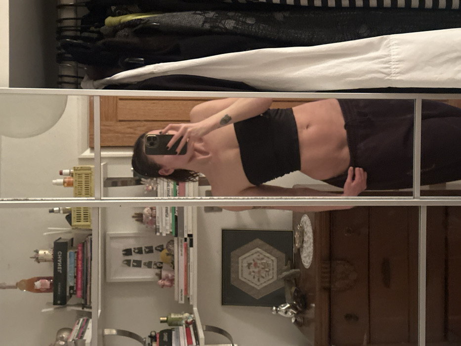 A mirrored image captures a person taking a selfie in a closet. They wear a black tube top and dark pants. The closet contains shelves filled with books, bottles, and decorative objects. Clothes hang above. A dresser is visible to the right of the person.