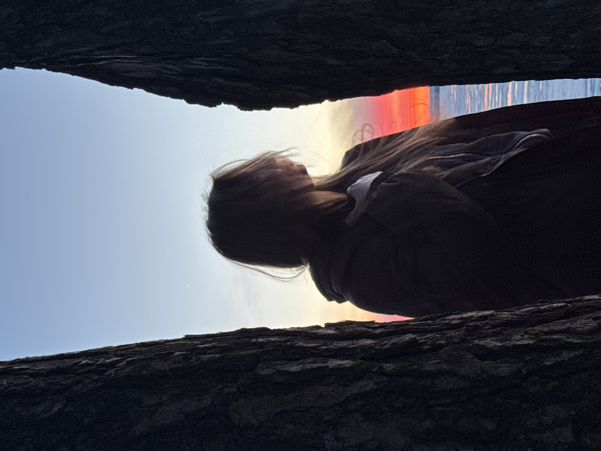 A person is silhouetted against a sky with vibrant colors, possibly at sunset, seen through a narrow gap between what appears to be tree branches or logs. The person has long hair, and is wearing a zippered jacket or sweater. The location is likely Chaikovsky, Russia, but without other confirming details, this is unverified.