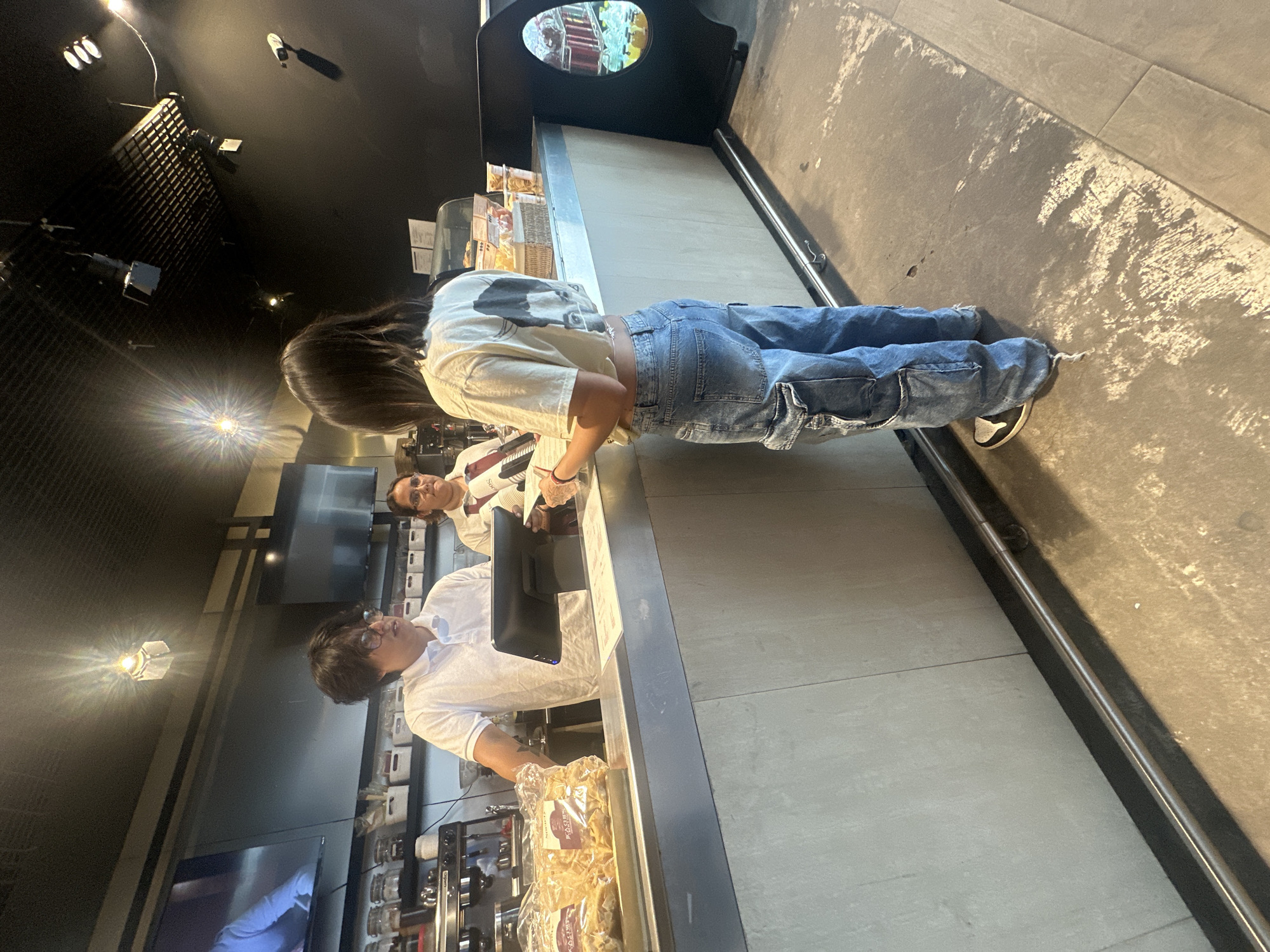 The image shows the interior of a cafe or bakery, likely in Mexico City, Mexico. A customer in light-colored jeans and a t-shirt stands at a counter. Two employees, one in glasses and a white polo shirt, and the other in glasses and a red and white striped shirt, are visible behind the counter. Food items are displayed under a glass or plastic covering. A cash register is positioned in front of the employees. Coffee makers and equipment are arranged behind them on the counter. The background shows a row of shelves containing containers or ingredients. Overhead, there is a black ceiling with visible lighting fixtures. The floor is tiled in gray.