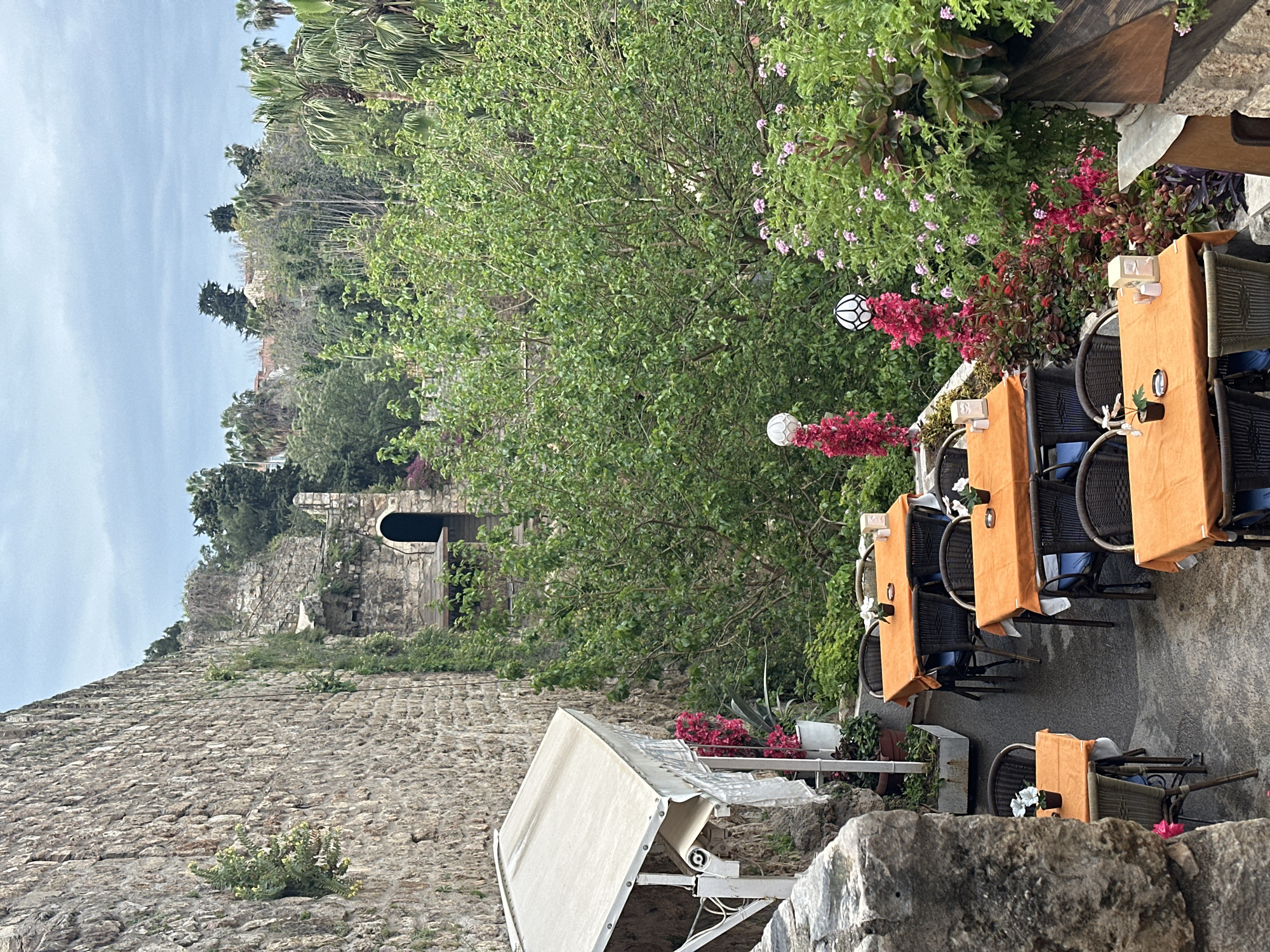 The image depicts an outdoor dining area in Antalya, Turkey, likely a restaurant patio. Tables are covered with orange cloths and set with flowers, chairs surrounding them. The area is bordered by stone walls and lush greenery, including flowering plants. In the background, there is a building with an arched doorway, suggesting historical architecture. The overall scene is one of a quiet, picturesque outdoor restaurant setting. The weather appears to be clear and sunny. No people are visible in the image.