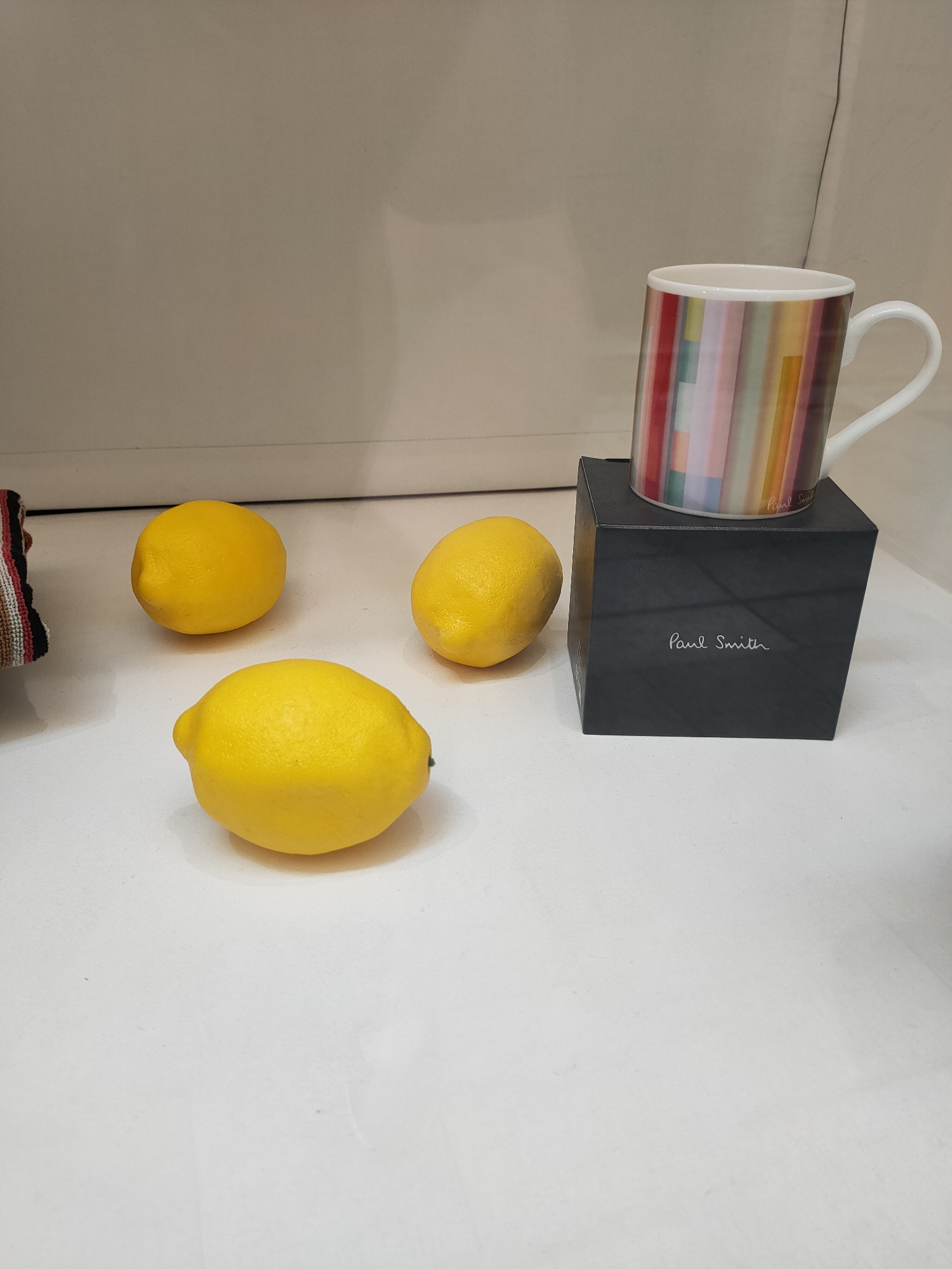The image depicts a still life arrangement on a white surface. Three lemons are present, two clustered together on the left and one slightly larger lemon in the foreground. To the right of the lemons, a black cubical box is visible, topped with a white mug. The mug has a handle and is decorated with vertical stripes of various colors, including red, yellow, green, and blue. The black box has "Paul Smith" printed in white script on its front. The background is a neutral, light-colored wall. There is no indication of the image being located in Perpignan, France.