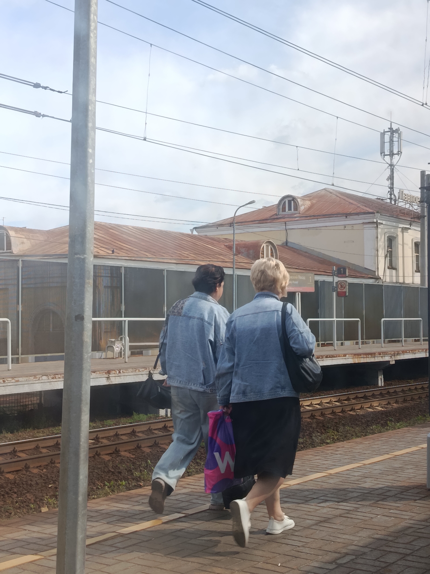 The image shows two individuals walking on a train platform in Pavlovsky Posad, Russia. The person on the left is wearing a denim jacket and light-colored pants, carrying a black bag. The person on the right is wearing a denim jacket and a black skirt, carrying a colorful shopping bag and a black handbag. They are walking away from the camera. In the background, there is a train station building with a rusty-colored roof and a telecommunication tower. The platform is paved with brick-like tiles and bordered by train tracks. The sky is cloudy.