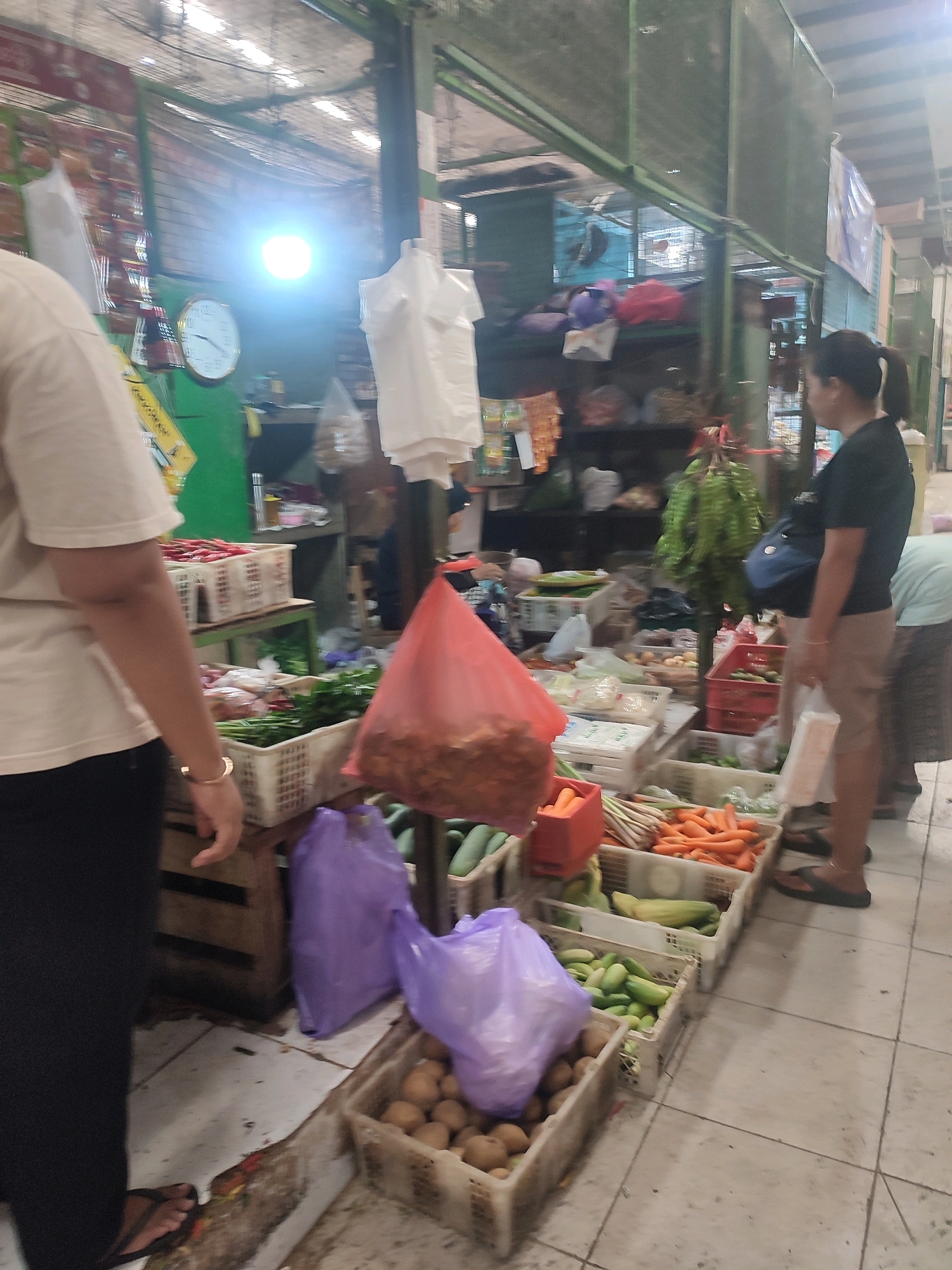 This image depicts a busy marketplace scene, likely in Yogyakarta, Indonesia, given the overall context. The foreground is dominated by a person from the waist down, wearing a light-colored t-shirt and black pants, with their left arm partially visible, displaying a bracelet. To the right, a woman with dark hair pulled back in a ponytail, wearing a black t-shirt and tan pants, is seen from the side, holding a white plastic bag. Behind her, another person is partially visible, wearing a light blue shirt and a patterned skirt.
The marketplace is filled with various fresh produce displayed in white and red plastic crates. Prominently featured are carrots, cucumbers, and what appear to be potatoes or similar root vegetables. There are also purple plastic bags, possibly containing groceries, placed among the produce. In the background, a clock with a white face and gold trim is visible on a green wall. Shelving units are stacked with more goods, and a bright light source illuminates the area, suggesting it's a covered market. The floor is tiled with a light gray pattern. The overall impression is one of a vibrant, working market environment.