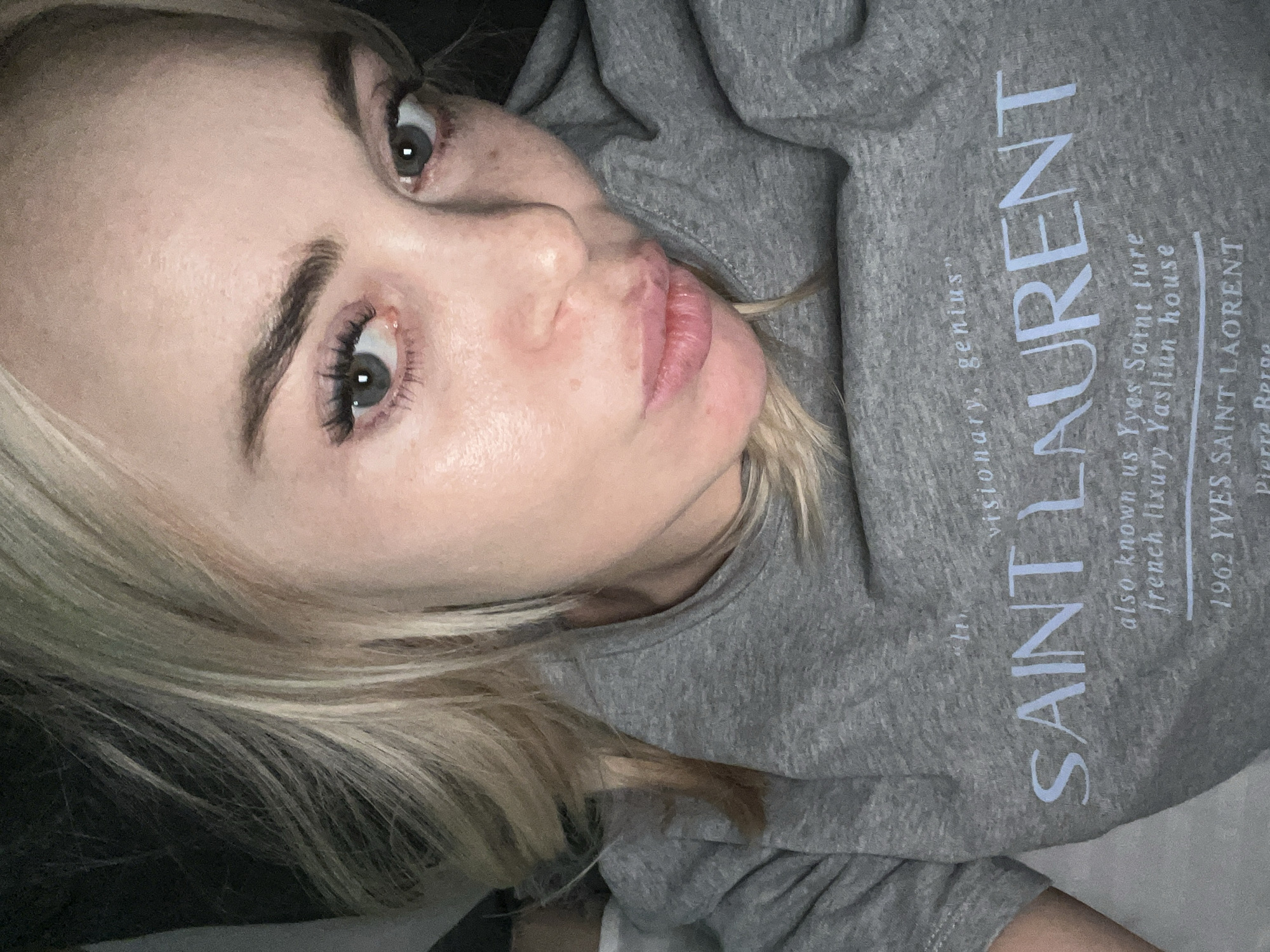 This media file is a close-up, low-angle shot of a young Caucasian woman's face. She has blonde hair, fair skin, and green eyes. Her expression is a playful pout, with her lips slightly parted. She is wearing a grey hooded sweatshirt with blue text that appears to be a brand logo. The text on the sweatshirt reads "SAINT LAURENT" in large, stylized letters, with smaller text below and to the left of the main logo. The smaller text includes phrases like "visionary, genius," "also known us Yves Saint ture," "french lixury Yasliun house," "1962 YVES SAINT LAORENT," and "Pierre Berge." The lighting is soft and appears to be natural, suggesting it may be daytime. The background is dark and blurred, indicating the focus is on the woman. The overall impression is one of a casual, intimate moment captured on camera.