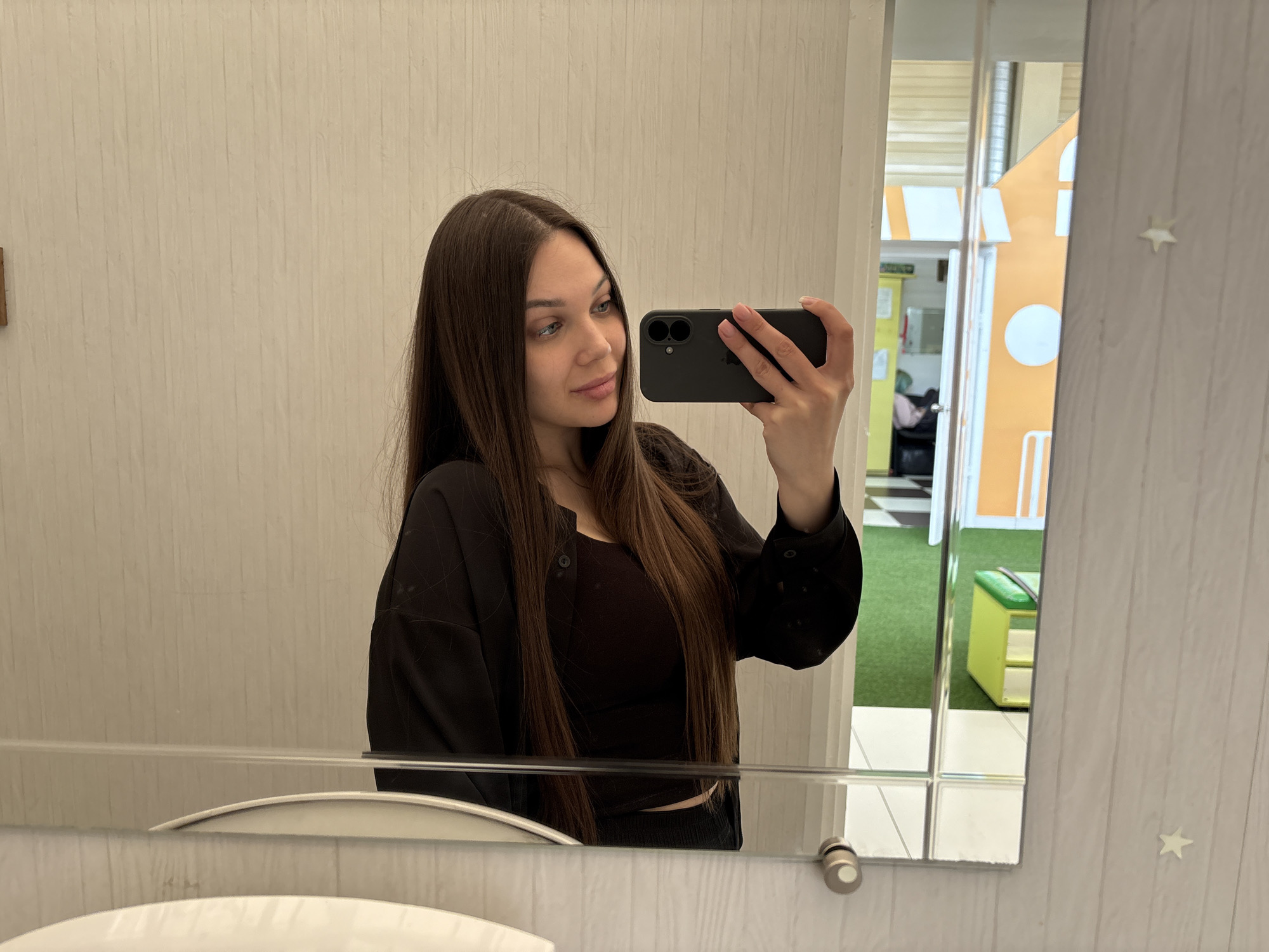 A young woman with long dark hair takes a selfie in a mirror. She holds a black smartphone in her right hand, her face slightly angled toward the camera. She wears a black top and a black button-down shirt. The mirror reflects part of a room with white walls and a striped pattern. Reflected in the mirror is also a play area with a yellow playhouse and green flooring, suggesting the location is likely a children's entertainment center. The photo was taken in the city of Chaikovsky, Russia. A white sink is partially visible at the bottom of the mirror.