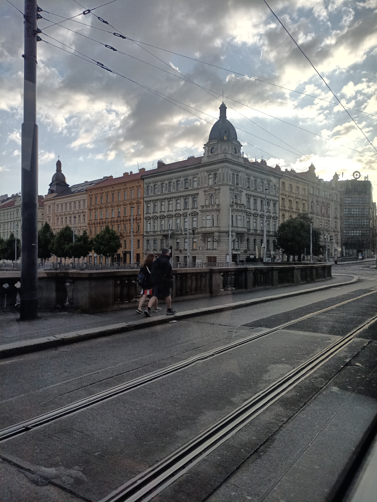 This image shows a street scene in Prague, Czechia, with tram tracks in the foreground and ornate buildings lining the street in the background. The sky is overcast with dramatic clouds. Two people are walking on the sidewalk in front of the buildings. One person is wearing a black jacket and shorts, and the other is wearing a black jacket and a striped skirt. They are walking away from the camera. The buildings appear to be a mix of architectural styles, with some featuring domed roofs and intricate detailing. The tram tracks are wet, suggesting recent rain. The overall mood of the image is somewhat somber due to the cloudy sky, but the architecture and presence of pedestrians suggest an active urban environment.