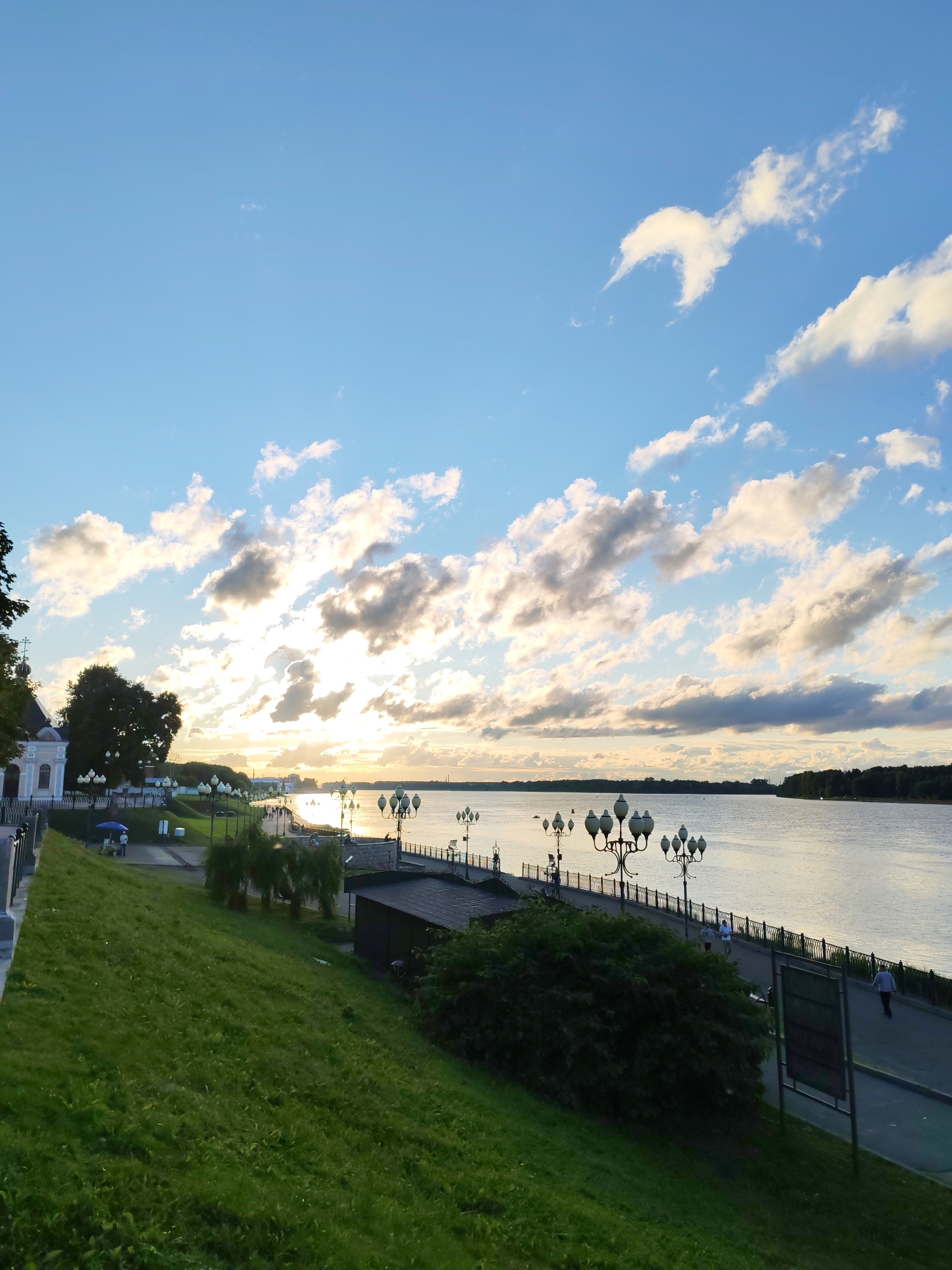 This media file depicts a scenic view of a riverfront promenade at sunset. The sky is a vibrant blue, adorned with scattered white and gray clouds, some of which are illuminated by the setting sun, casting a warm glow. The sun itself is visible near the horizon, creating a shimmering reflection on the calm water of the wide river.
The scene shows a paved promenade lined with decorative lampposts, suggesting a public space designed for leisurely strolls. A dark-railed fence separates the promenade from a grassy, sloping hill in the foreground. On the left side of the promenade, a white building with a dome and a cross can be seen, possibly a church or a historical structure. A few trees are scattered along the embankment, with a large, dark tree dominating a portion of the left side.
Several people are visible on the promenade, walking and enjoying the evening atmosphere. One person is running, and another is standing near a blue umbrella. The overall mood of the image is peaceful and serene, characteristic of a pleasant evening by the water. The time of day is clearly indicated by the low angle of the sun and the warm, diffused light. The lush green grass on the hill suggests a temperate climate. The location context of Bol'shie Mkhi, Russia, provides a specific geographical setting for this tranquil riverside scene. No visible text is present in the image.