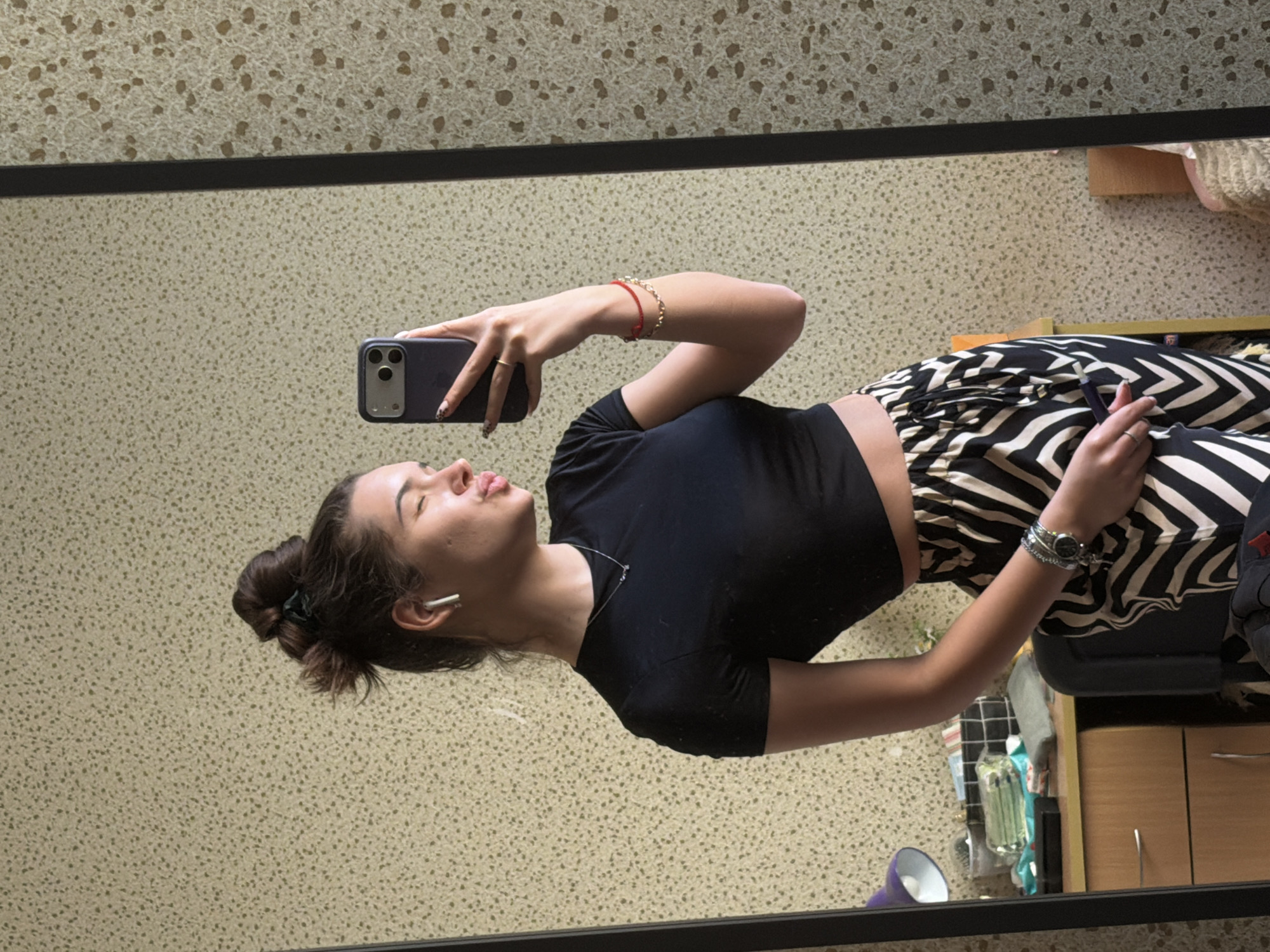 A young woman is reflected in a mirror while taking a selfie with a smartphone. She is wearing a black crop top and zebra-striped pants. She has her hair up in a bun and is wearing an earpiece. Bracelets adorn her wrists, and a watch is visible on her left wrist. The background includes a patterned wall, a dresser with drawers, a purple lamp, and other household items. There is no evidence to suggest that the image was taken in Sofia, Bulgaria.