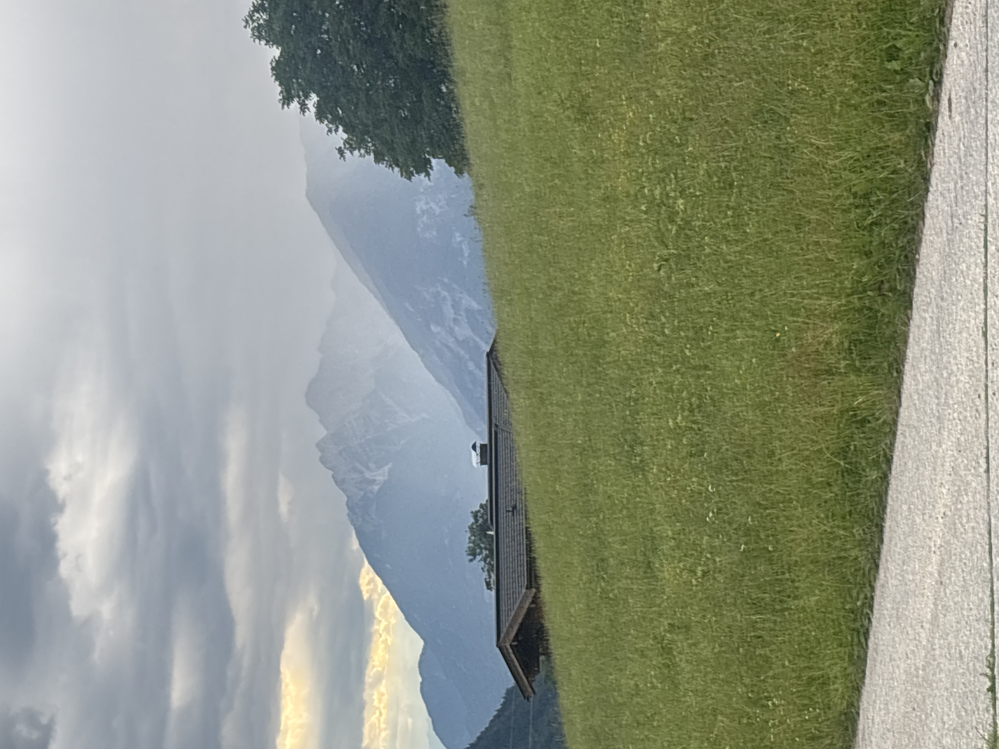 A wooden cabin with a dark, shingled roof and a chimney is situated on a grassy hillside. The cabin is partially obscured by the foreground, which is a vibrant green meadow dotted with small white flowers. In the background, majestic mountains rise, their peaks shrouded in mist and clouds. The sky is overcast, with patches of sunlight breaking through the clouds on the left side of the image, casting a golden glow. A paved path or road runs along the bottom edge of the frame. The scene evokes a sense of natural beauty and tranquility in a mountainous region. The location is Hyna, Austria.
