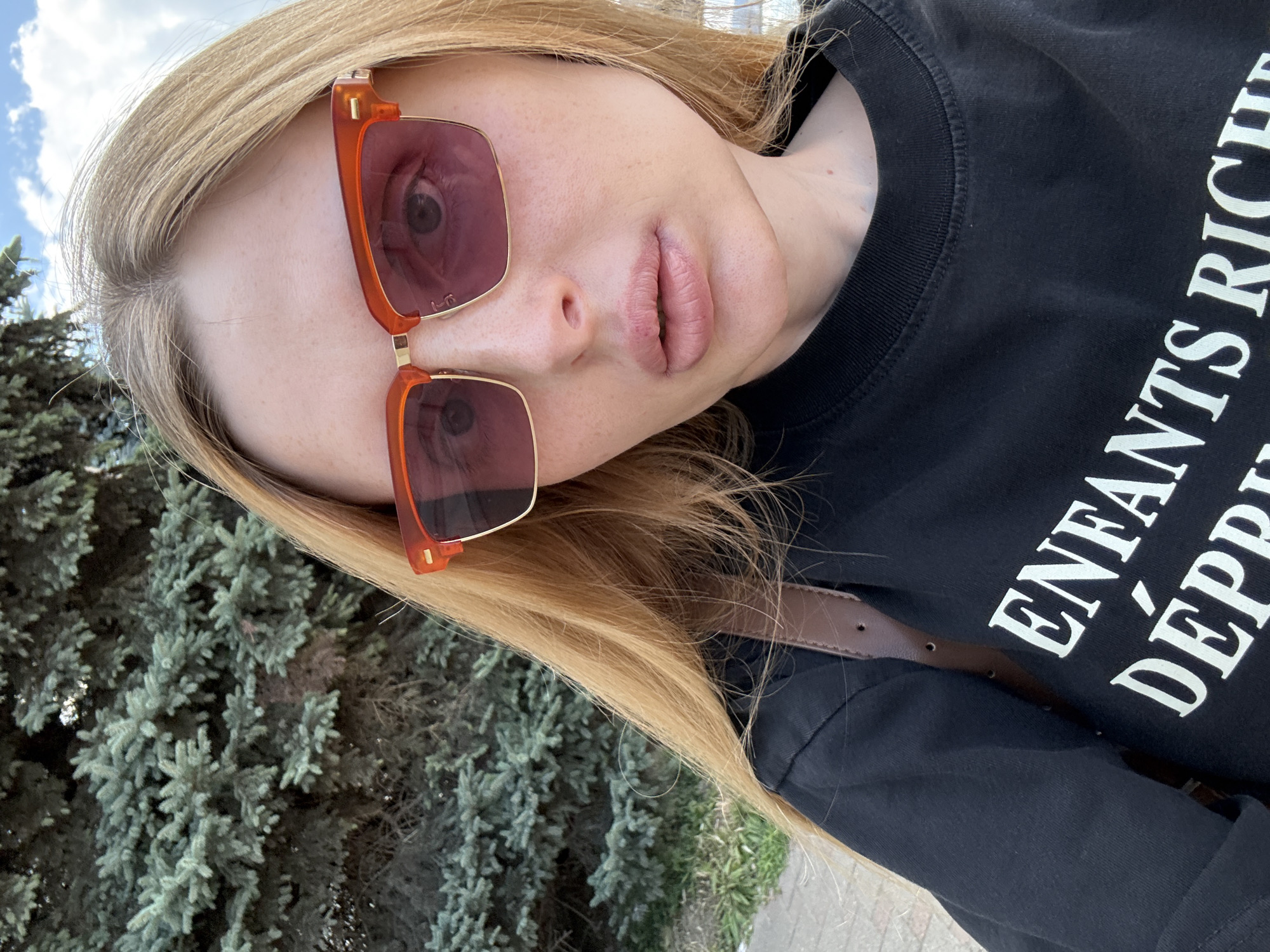 A close-up, outdoor, waist-up selfie shows a young woman with blonde hair and fair skin. She is wearing oversized sunglasses with orange frames and pink lenses, and a black t-shirt with white lettering that reads "ENFANTS RICH DÉPR". She has full lips and freckles across her cheeks and nose. Behind her is a large evergreen tree. The background also shows a patch of sidewalk and a cloudy sky. The scene appears to be in an urban park or residential area.