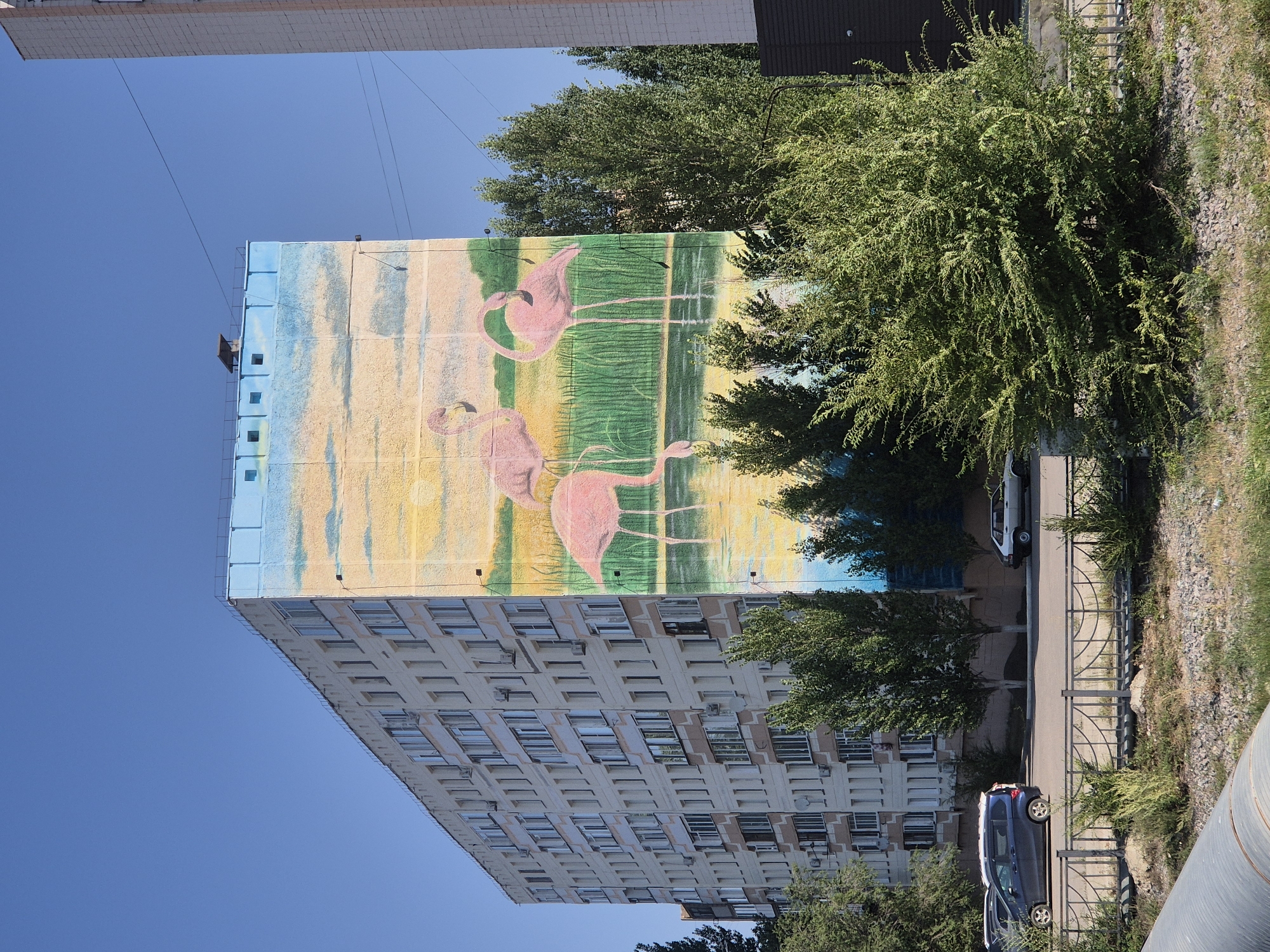 The image shows a large apartment building in Kokshetau, Kazakhstan, with a mural painted on its side. The mural depicts three pink flamingos standing in water with reeds. The building is multi-story, with many windows and balconies. In front of the building, there is a street with parked cars and trees. The sky is clear blue. The mural appears to be a prominent feature of the building's exterior.