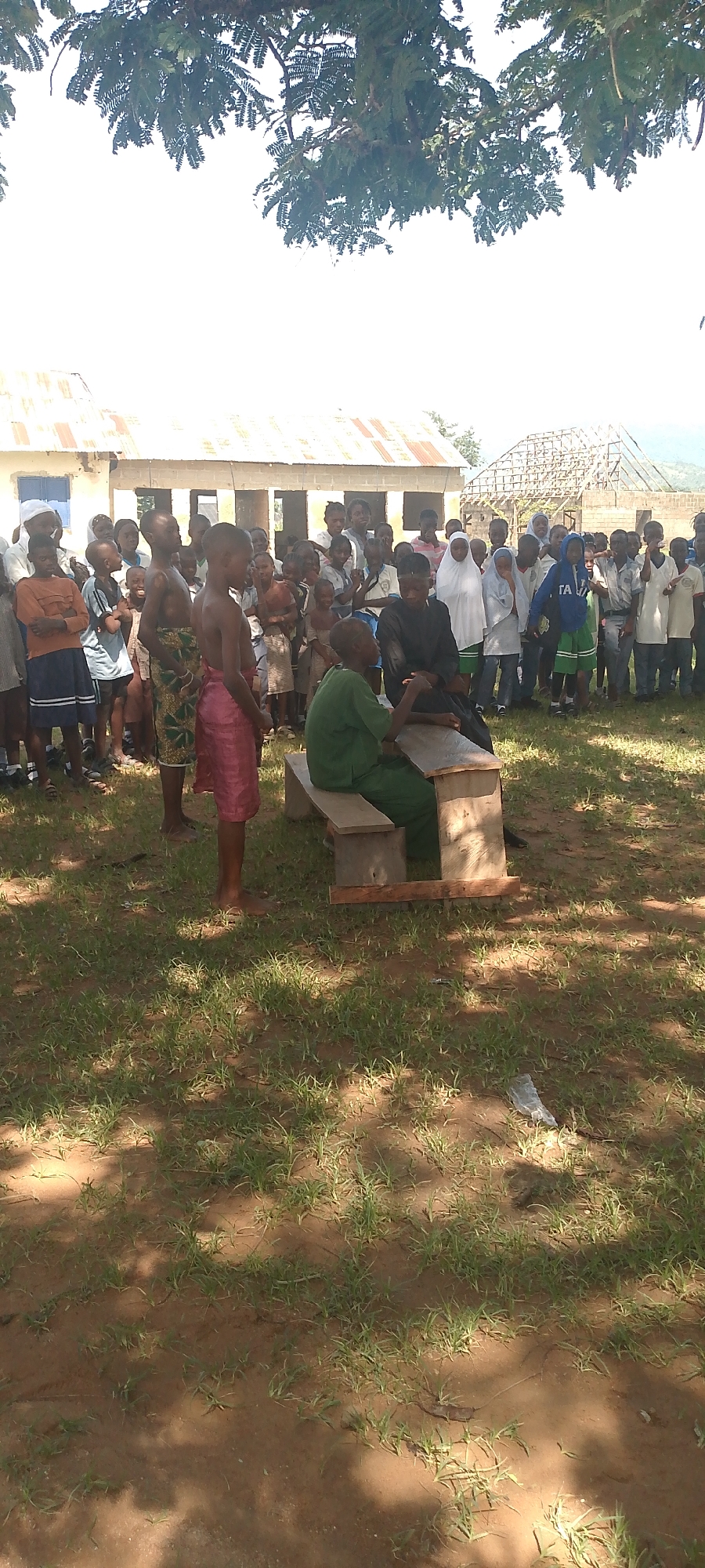 In a schoolyard, a group of children and adults are gathered outdoors, some standing and others seated. A few children, appearing to be school-aged boys, are in the foreground. One boy wears a pink cloth wrapped around his waist, while another is shirtless and wears a patterned cloth. A young boy in a green garment is seated on a wooden bench, facing away from the camera, and is in conversation with a man in a dark outfit, also seated on the bench. Many children are looking towards the seated pair. Behind the seated individuals, a larger group of children and some adults are assembled, some wearing hijabs. The environment appears to be a rural or semi-rural setting with buildings that seem to be part of a school. One building has a corrugated metal roof, and another in the background is under construction. The ground is covered in grass and dirt, with shadows suggesting the presence of trees overhead.