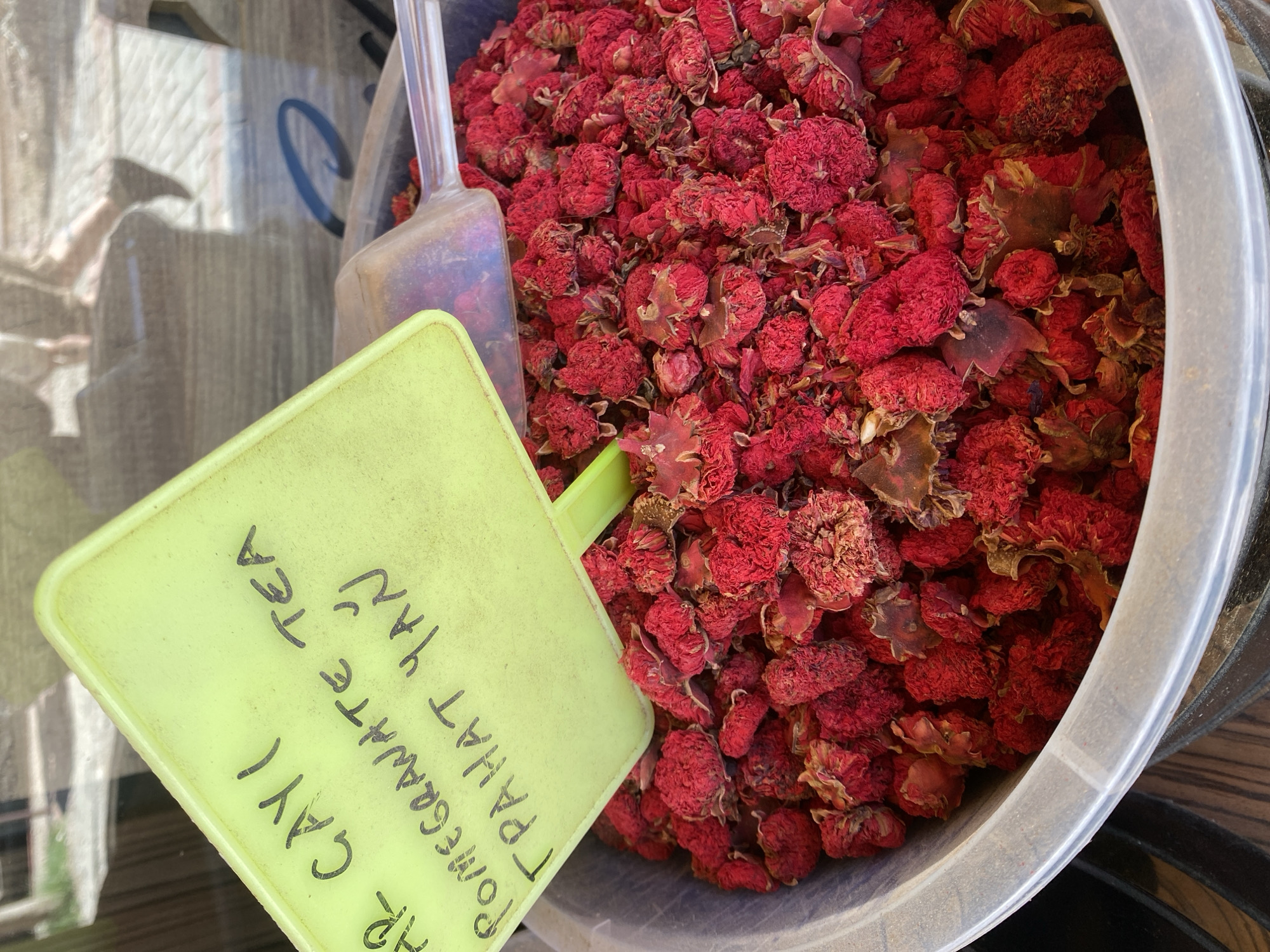 An eye-level, medium shot reveals a plastic tub filled with dried, reddish-purple pomegranate blossoms for tea, accompanied by a clear plastic scoop and a handwritten, yellow sign. The tub takes up a majority of the frame, filled to the top with the tightly packed dried blossoms, some with remnants of brown leaves attached. A clear plastic scoop is partially submerged in the blossoms. To the left of the tub, a yellow sign with handwritten text in black ink reads "AR CAYI / POMEGRANATE TEA / TRAHAT YAN."
The plastic tub is off-white, and part of a dark object is visible at the lower right of the frame. The environment appears to be a marketplace, with blurred reflections visible in the upper left. The photograph was likely taken in Alanya, Turkey, based on the use of the Turkish word "çay" for tea and "nar" for pomegranate.