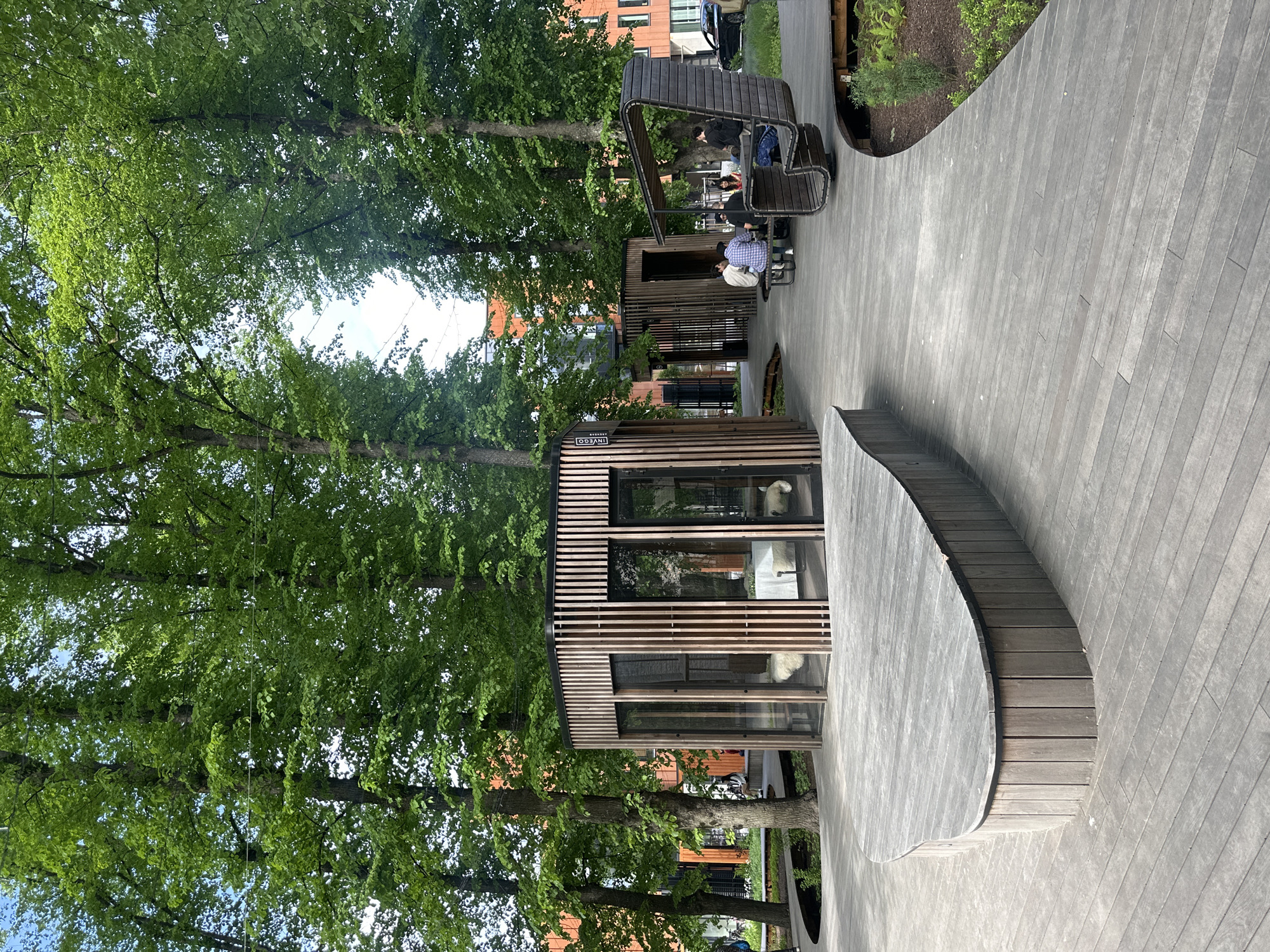 This image depicts a modern outdoor seating area in Tallinn, Estonia, likely within a contemporary urban development. The scene features a curved wooden boardwalk with several distinct structures and seating arrangements.
In the foreground, a light grey wooden boardwalk curves gently to the right. The boardwalk is constructed from evenly spaced planks and has a raised, curved edge made of vertical wooden slats on the left side.
Further into the scene, a small, contemporary pavilion with a slatted wooden exterior and glass walls is visible. Inside, a white table with chairs is set up, and two white sheepskin rugs are placed on the floor. Adjacent to this pavilion is another similar structure.
To the right of the pavilions, a unique, dark wood, sculptural bench is situated. This bench has a high, curved back and appears designed to seat multiple people. A group of four individuals is seated at a table in front of this bench, engaged in conversation.
In the background, modern buildings with orange facades and large windows are visible through the lush green foliage of tall trees. The trees are abundant, with dense green leaves, suggesting a spring or summer setting. The sky is visible through the canopy, appearing overcast or partially cloudy. A dark-colored car is parked on the street in the far background. The overall impression is a blend of modern architecture and natural elements within an urban environment.