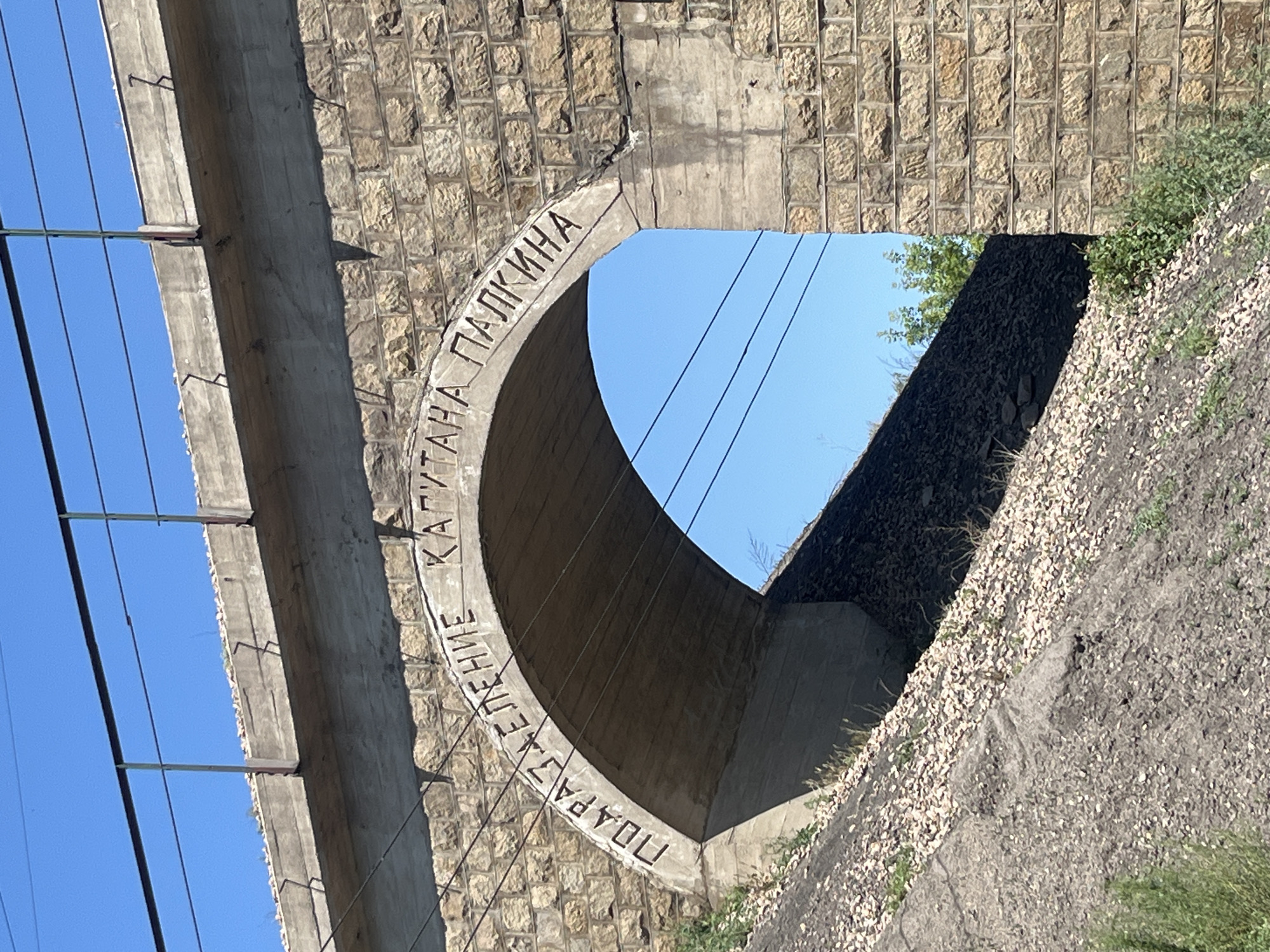 The image shows an arched underpass, likely part of a railway bridge, constructed with rough-hewn stone blocks on the exterior and a smooth concrete interior. Carved into the concrete arch is text in Cyrillic script, reading "ПОДРАЗДЕЛЕНИЕ КАПИТАНА ПАЛКИНА". This translates to "Captain Palkin's Unit". The underpass is situated on a slope covered with loose gravel and vegetation, with power lines visible above the arch and against a clear blue sky. The overall scene suggests an infrastructure element in a somewhat rural or industrial setting. The provided information about the city of Aqtau, Kazakhstan, is not directly verifiable from the visual elements alone.