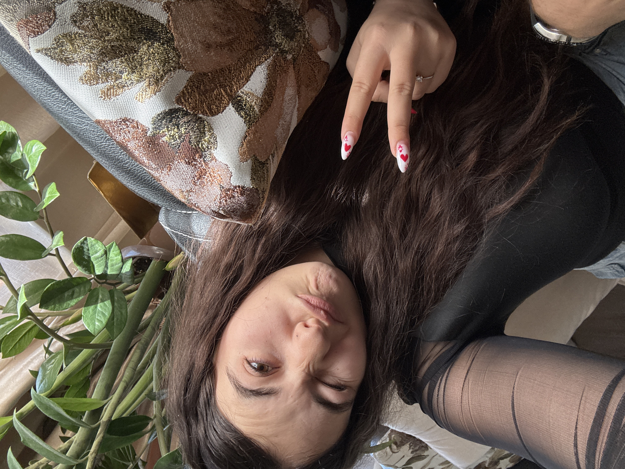 A young woman with long brown hair and one eye closed is the central subject of the image. She is wearing black attire and dark-colored tights. Her nails are painted white with red heart designs. She is making a "peace" sign with her fingers. A patterned pillow rests next to her head, and a potted plant with green leaves is situated in the lower left corner of the frame. Based on the background elements, it's plausible the photo was taken indoors, possibly in a home setting. However, there is no information present that definitively places the scene in Plovdiv, Bulgaria, or any other specific location.