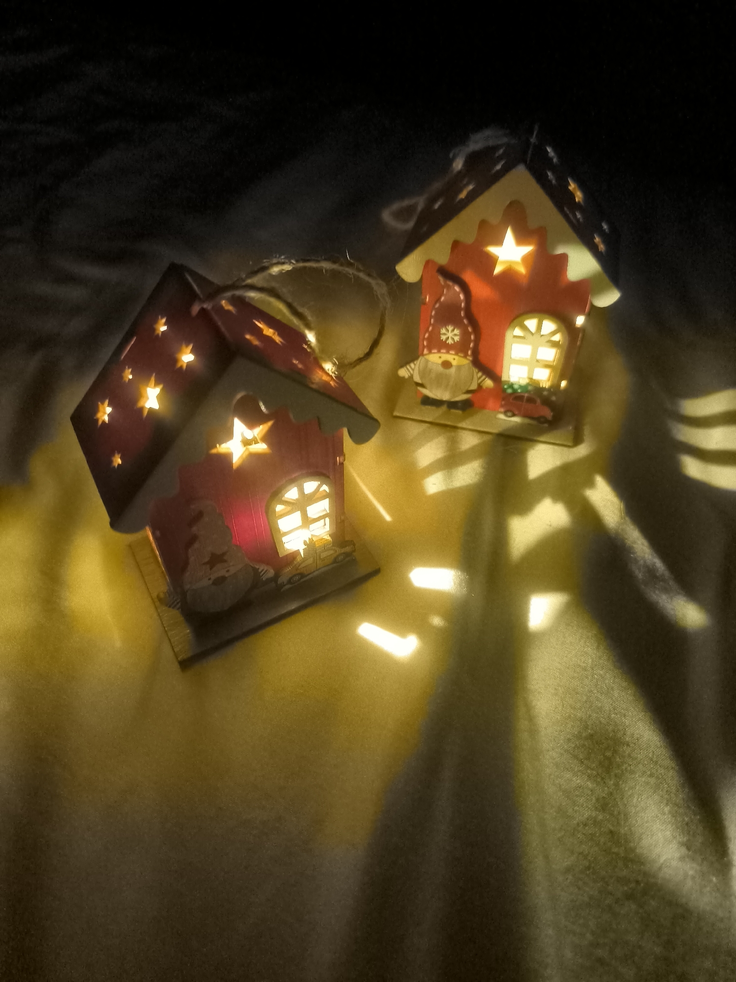 Two decorative, illuminated Christmas houses are placed on a textured, yellow surface, likely a blanket or cloth, under dim lighting. The house on the left is red with yellow stars cut out, some of which are illuminated. It features a cutout of a gnome on the front. The house on the right is also red and yellow, with a cutout of a gnome wearing a red hat with a snowflake and a small red car in front of it. Both houses have illuminated windows. The lighting creates stark shadows and bright spots on the surface. The image does not depict any elements that would indicate its location in Horishni Plavni, Ukraine.