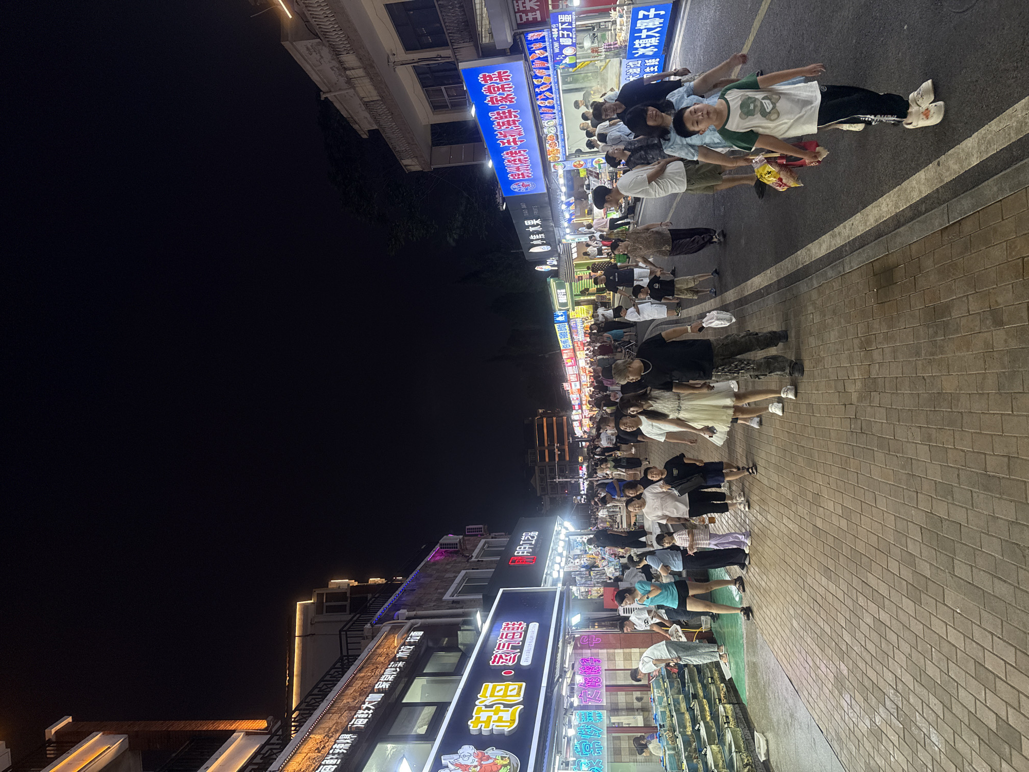 This image depicts a bustling night market scene in Qinhuangdao, China. A street lined with numerous food stalls and shops is filled with people. The stalls display a variety of goods, including seafood in tanks, as well as illuminated signs advertising food items in Chinese characters. Pedestrians, including families with children, are walking along the paved walkway and the street. Some individuals are engaged in browsing, while others are walking or standing. The architecture suggests a commercial district, with multi-story buildings on either side of the street. The lighting is dominated by the vibrant neon signs of the stalls and the streetlights, creating a lively nighttime atmosphere.