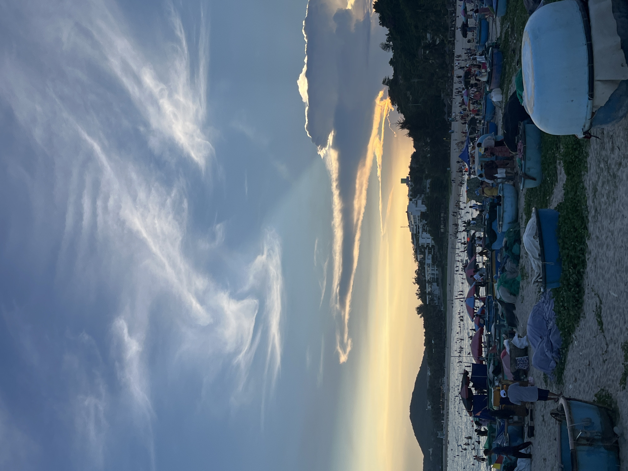 The image depicts a beach scene in Phu Thuy Ward, Vietnam, during what appears to be late afternoon or early evening. The sky is a gradient of blue, with streaks of wispy white clouds and dramatic, illuminated cumulus clouds on the right. A warm, golden light suggests the setting sun is near the horizon, casting a soft glow over the landscape.
The beach is populated with numerous people, many of whom are in the shallow water, swimming or wading. Along the shoreline, there are many traditional Vietnamese fishing boats, primarily round basket boats, some of which are stacked or covered with tarps. Umbrellas are set up on the sand, providing shade for beachgoers.
In the background, a hill covered with lush vegetation is visible, with buildings and structures scattered across its slopes, indicating a coastal town or resort area. The overall atmosphere is one of a busy, recreational beach with signs of local fishing activity.