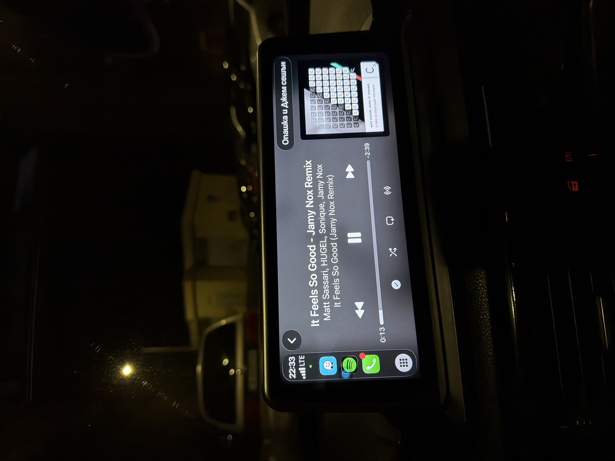 The image displays a car's interior at 22:33, focused on an illuminated infotainment screen showcasing the Apple CarPlay interface with the Spotify application. The screen shows the track "It Feels So Good - Jamy Nox Remix" by Matt Sassari, HUGEL, Sonique, Jamy Nox currently playing, with 0:13 elapsed and -2:39 remaining. An LTE connection and various application icons (Messages, Spotify, Phone) are visible on the left side of the screen. The right portion of the screen displays Cyrillic text "Опашка и Джем сешън" (Queue and Jam Session) above a grid-like visualizer composed mainly of repeating 'C' characters, with some green and red highlights. Below the screen, portions of the dark dashboard are visible, including illuminated red indicators for "DTC" and a window control.
Outside the vehicle, a nighttime urban environment in Varna, Bulgaria, is visible through the windshield. A bright yellow streetlamp is prominently positioned in the upper left background, illuminating a section of the dark scene and creating a glare on the windshield, indicating minor condensation or residue. The rear section of a light-colored vehicle is partially discernible directly behind the infotainment unit, and additional blurred vehicle shapes and ambient lights are present in the distant background. A faint, light-colored building can be seen on the far left of the background. The scene suggests a stationary vehicle in an urban or parking area after dark.