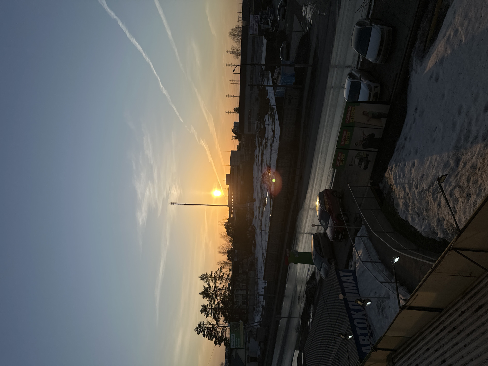 The image captures a sunset view in what appears to be the city of Chaikovsky, Russia. The sun is low on the horizon, casting a bright orange glow that contrasts with the blue sky and contrails left by airplanes. Below the horizon, there is an industrial area, possibly a parking lot or a business district.
A variety of objects are apparent in the image: power lines, trees, roads, cars, and a commercial building marked with the word "АВТОКЛИМАТ" and text in Russian, indicating the presence of a store or a business dealing with automotive climate control systems. Adjacent to the building, there are advertisements or billboards.
The scene suggests a cold climate as there are patches of snow visible on the ground. The presence of cars and buildings points to an urban setting, and the setting sun suggests it's either early morning or late afternoon.