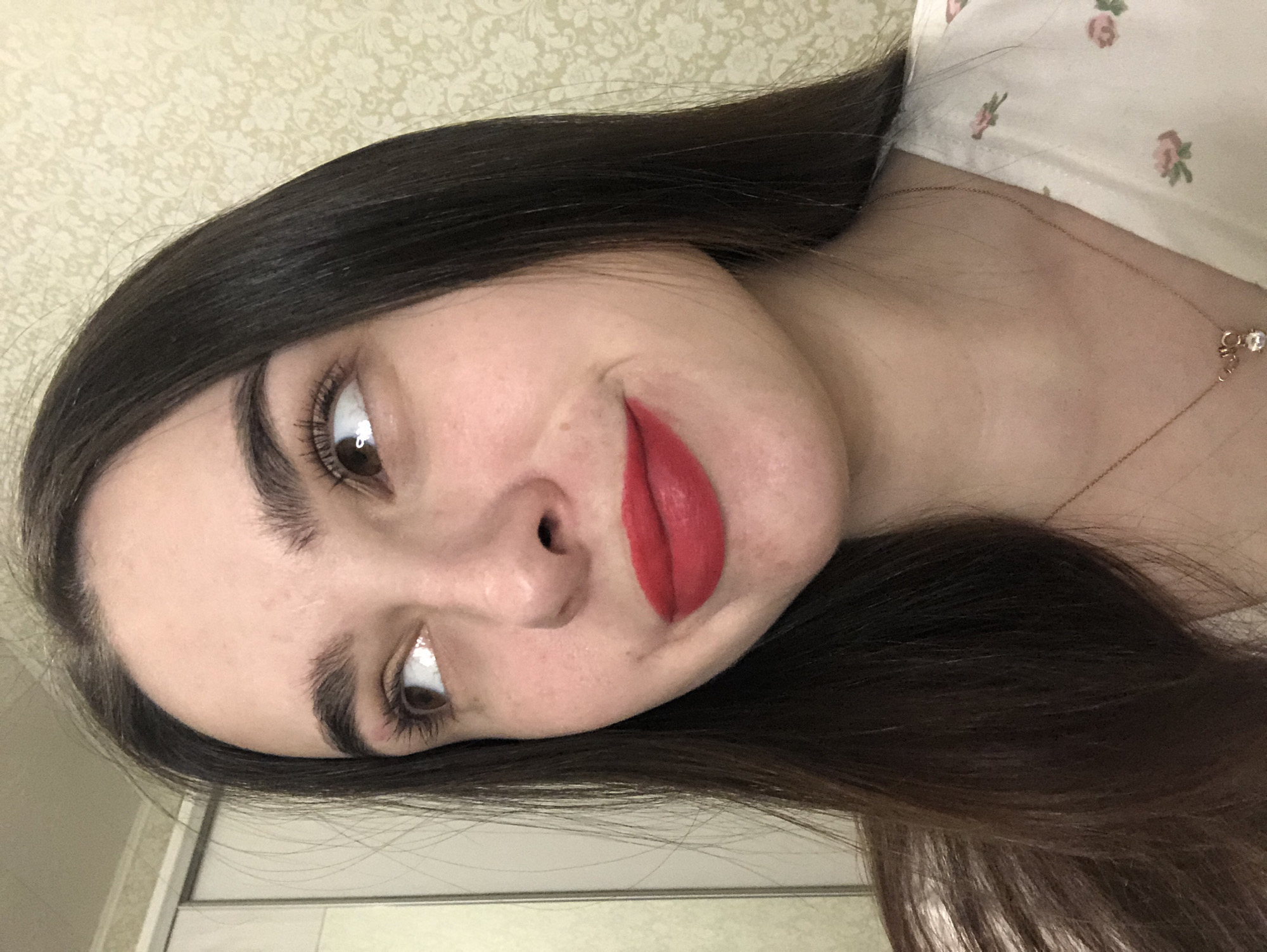 A woman with long, dark hair is looking at the camera. She has fair skin, dark eyebrows, and is wearing red lipstick. She is wearing a light-colored top with a floral pattern and a gold necklace. The background is a beige wall with a faint pattern. A white door or cabinet is visible in the lower left corner. The image appears to be taken indoors.
The location in the city of Noviy, Russia is not discernible from the image.