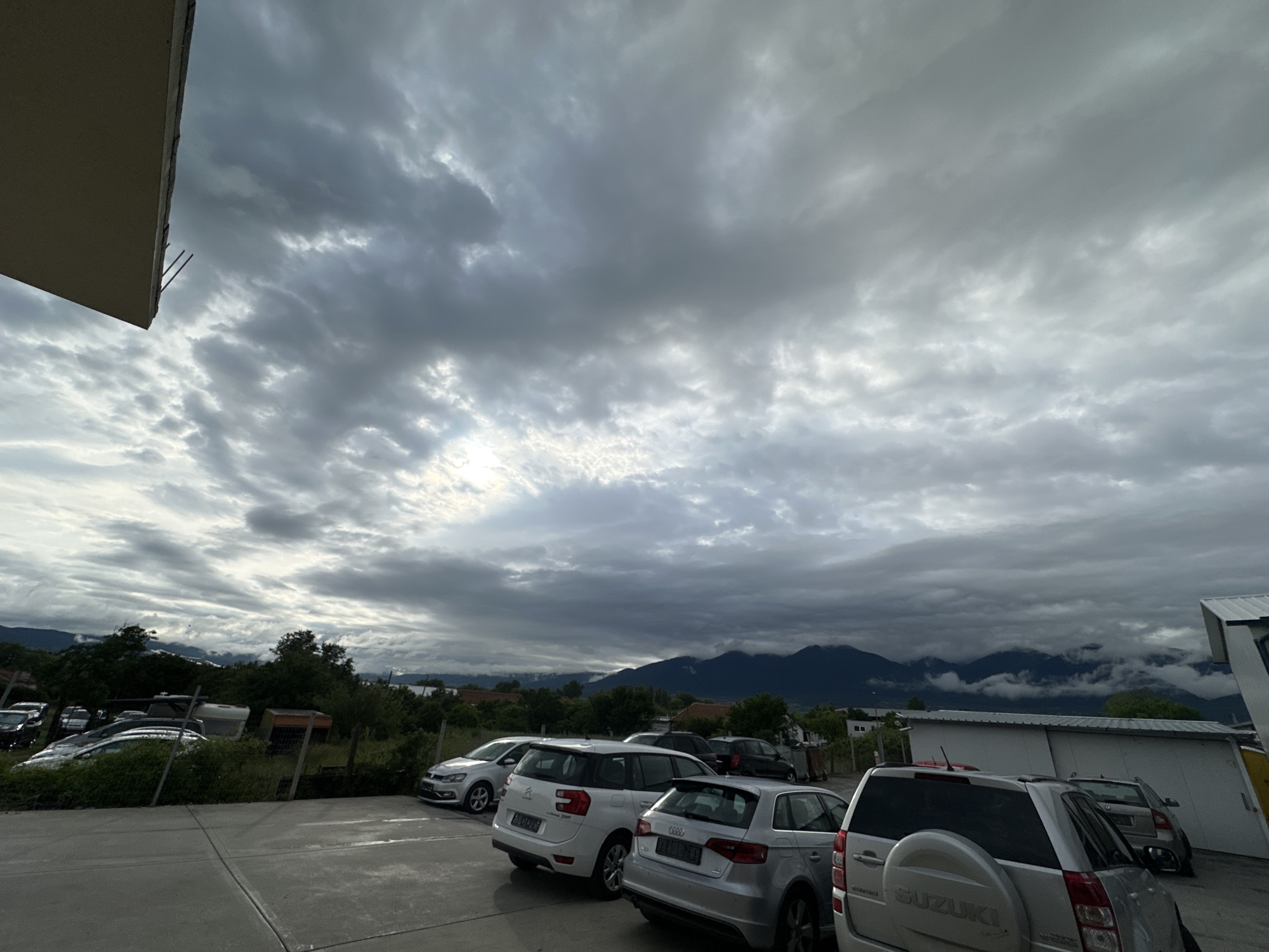 A cloudy, overcast sky looms above a parking lot filled with several vehicles. The scene appears to be in Piperevo, Bulgaria, as indicated by the presence of cars, a building, and mountainous terrain in the background. Among the vehicles are a white Citroën minivan, a silver Audi hatchback, and a silver Suzuki SUV with a spare tire mounted on the rear. The parking lot is paved, and a sidewalk is visible in the foreground. In the background, lush green trees and vegetation surround some of the parked vehicles, and further back, mountains are shrouded in mist and clouds. A portion of a light-colored building with exposed rebar is visible in the upper left corner of the image.