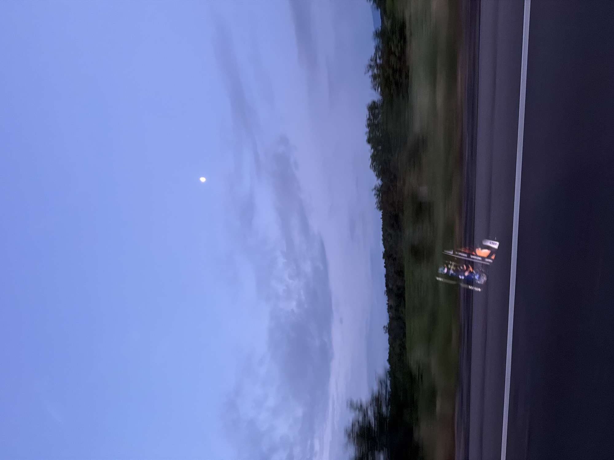 This media file presents a dynamic view from a moving vehicle, likely a car, over a road near Kalugerovo, Bulgaria, during twilight. The sky dominates the upper and left portions of the frame, displaying a serene palette of light blue and purplish hues, indicative of either dawn or dusk. Scattered, wispy clouds are visible, and a distinct crescent moon shines brightly in the upper left.
Adjacent to the road on the left, a blurred treeline or dense vegetation suggests passage through a forested or rural area, emphasizing the motion of the capturing vehicle. The right side of the image features a dark asphalt multi-lane road, clearly marked by a white solid line on its outer edge and a faint, blurred white broken line separating lanes.
A go-kart or a similar small racing vehicle is visible on the road in the mid-right. It features an orange and black color scheme with blue elements, reminiscent of racing liveries. Text that appears to say "KART" is discernable on its side. The go-kart itself is somewhat blurred, contributing to the overall sense of motion captured in the image. The entire scene, from the blurred trees to the moving go-kart and the slightly indistinct road markings, conveys the feeling of being in transit.