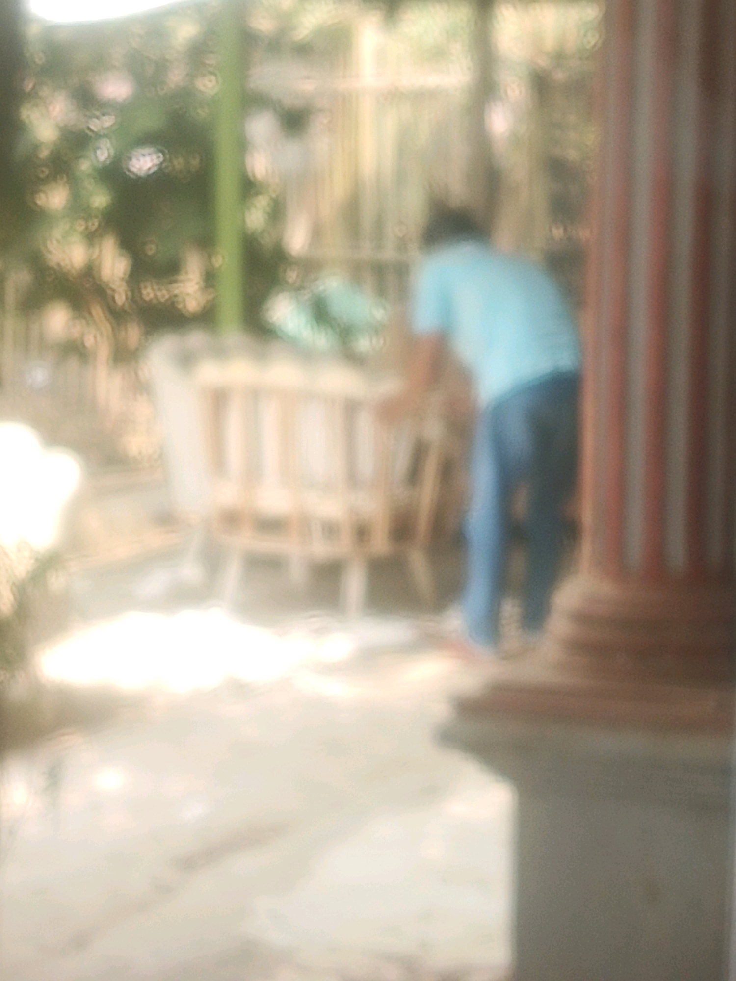 The image shows a blurred scene, likely taken outdoors in a residential area of Sidamukti, Indonesia. In the center, a person is bent over, seemingly attending to a wooden bassinet or cradle. The person is wearing a light blue t-shirt and blue trousers. To the right, a large, striped column, possibly part of a building or porch, is visible. The background is out of focus, but appears to include vegetation and some fencing, suggesting a garden or yard setting. The ground in the foreground is paved, with some bright highlights indicating sunlight. The overall impression is that of a candid, possibly domestic, moment captured in an everyday environment.