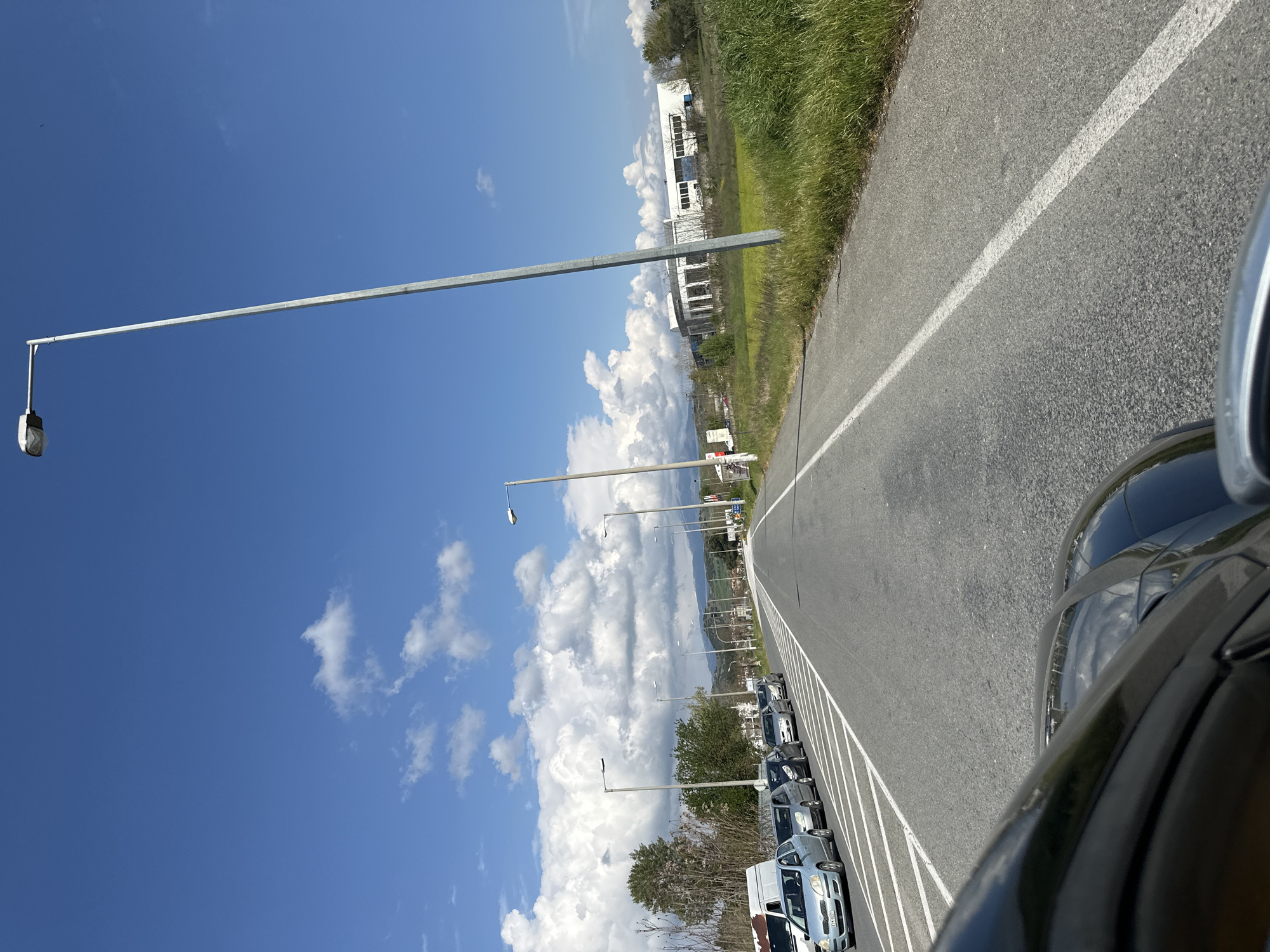 A road in Lefkonas, Greece is lined with parked cars, stretching into the distance under a blue sky with puffy clouds. Streetlights line the road. The vegetation borders the road on one side. A building is visible in the background. The sky is clear, and the road surface appears to be asphalt.
