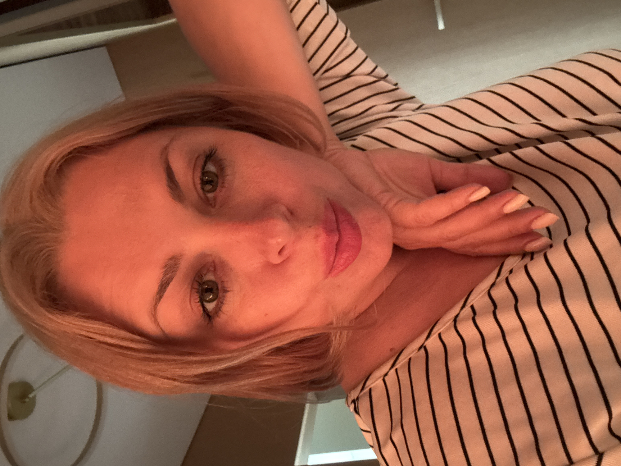 A woman with blonde hair and green eyes is captured in a selfie, her face turned towards the camera. She is wearing a black and white striped shirt and her left hand is resting on her chin, showcasing her peach-colored fingernails. The lighting in the image suggests it might be evening or indoors, with a warm, artificial glow illuminating her face. The background is blurred, but hints of a domestic setting can be seen, possibly a home. The woman's expression is neutral, with a slight hint of a smile. There is no visible text in the image.