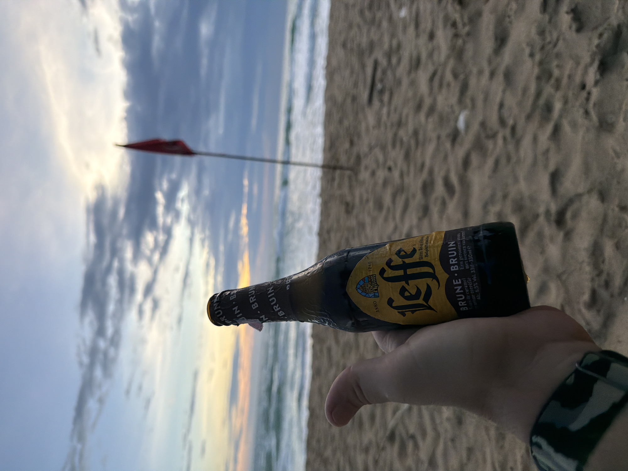 A person's hand holds a brown glass bottle of Leffe Brune beer on a sandy beach. The bottle has a yellow label with the Leffe logo and text indicating "BRUNE - BRUIN." Behind the bottle, the ocean with waves is visible, and further back, a cloudy sky with the sun setting can be seen. A red flag on a pole stands in the sand to the right of the bottle. The image appears to be taken at sunset on a beach in Choeng Thale, Thailand.