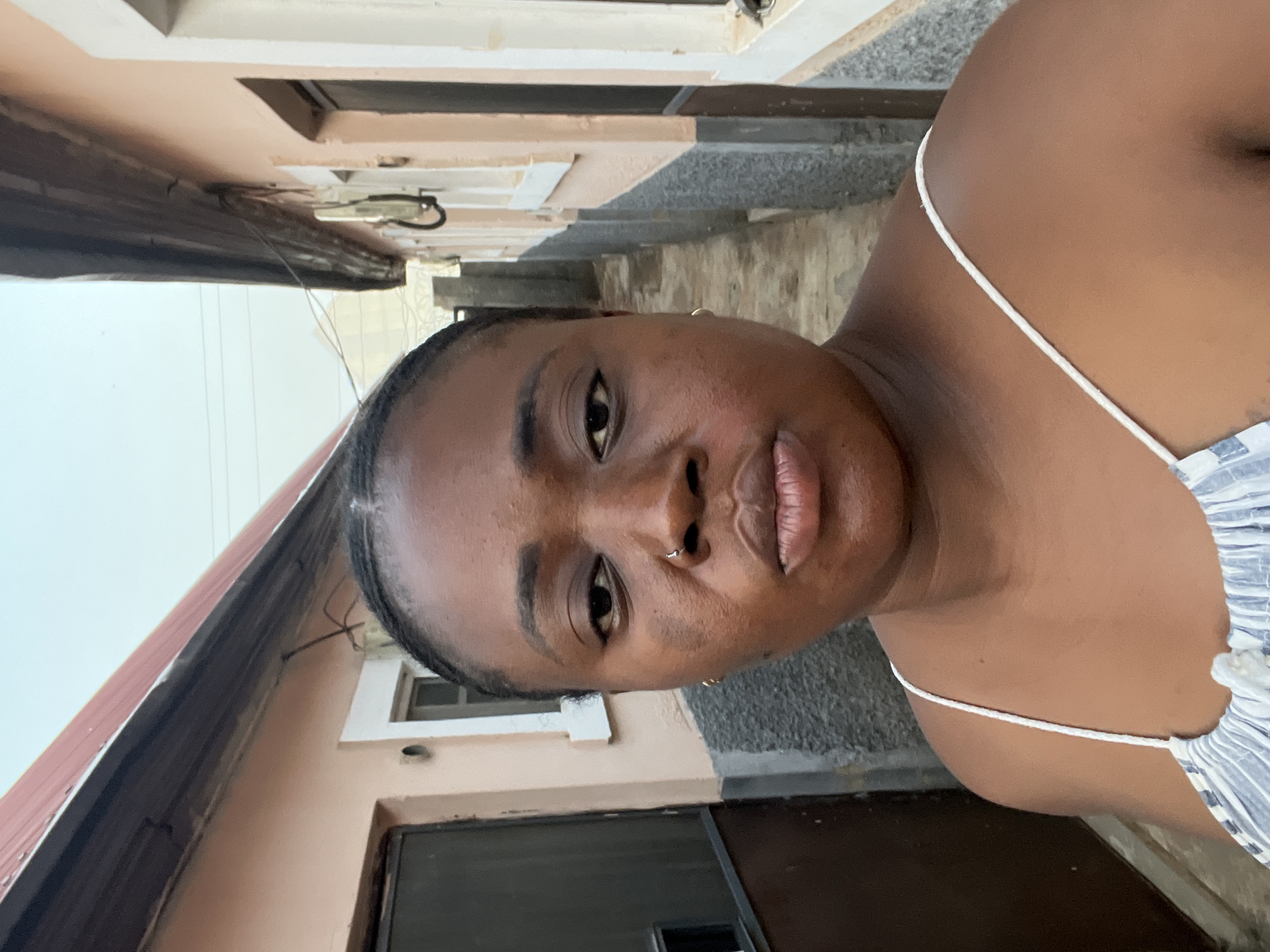 A young, dark-skinned woman is centered in this close-up, vertical selfie. She has a nose ring in her left nostril and a small hoop earring in her left ear. She is wearing a white spaghetti-strap top.
Behind her is a building. The lower portion is dark with what appears to be a door. The upper portion of the building is light beige or peach, with white trim around a window. A section of roof is visible at the left edge of the frame, featuring a reddish-brown hue. The image seems to be taken outdoors, potentially in an alleyway or narrow street. The setting is consistent with a residential area in Abuja, Nigeria, indicated by the architectural style and environment.
