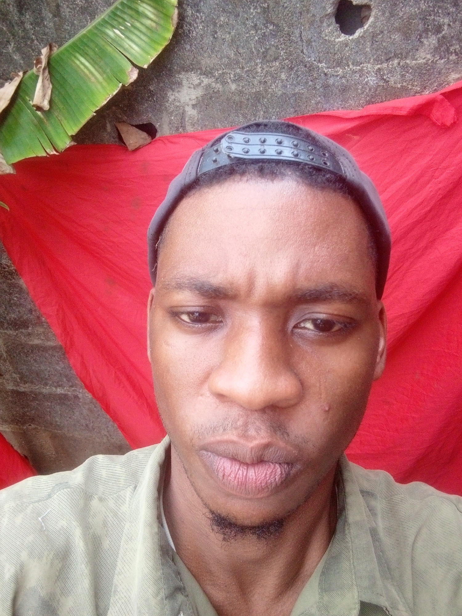 A medium shot depicts an adult male of African descent, with short hair covered by a dark head covering. His features include dark eyes, a broad nose, and a sparse goatee. He is wearing an olive-green collared shirt. The background is a juxtaposition of natural and artificial elements. A large green banana leaf extends from the top left, contrasting with a textured gray concrete wall. A vibrant red cloth is draped behind the subject. The image appears to be taken in Unknown, Unknown.