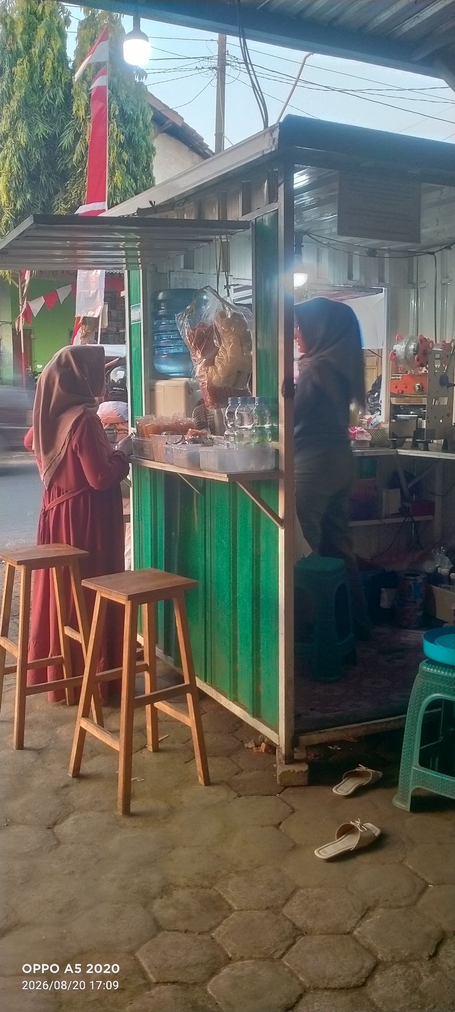 A street food stall is depicted in the city of Kedung, Indonesia. Two women wearing hijabs are at the stall. The woman on the left is wearing a maroon dress and is standing in front of the counter. The woman on the right is wearing a dark hijab and is inside the stall. The stall has a green corrugated metal exterior and a wooden counter displaying various food items. Behind the counter, there is a large water dispenser and bags of snacks. In front of the stall, there are two wooden stools and a green plastic stool inside the stall. The ground is paved with hexagonal concrete blocks. A red and white Indonesian flag is visible to the left of the stall, and trees are in the background. Two pairs of sandals are on the ground in front of the stall. The timestamp on the image indicates August 20, 2026, at 17:09.