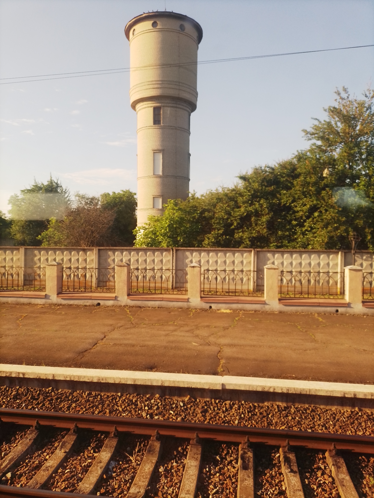 An outdoor, slightly hazy, horizontal medium shot captures a water tower and a train track in Pokrovske, Ukraine. The water tower, made of light-colored bricks, has a cylindrical shape with several distinct bands and two small rectangular windows. It is positioned on the left side of the image and rises above a dense line of green trees. In front of the trees, a concrete fence with a repeating diamond pattern is visible, supported by concrete pillars and a black metal railing. A cracked asphalt surface is in front of the fence. The foreground is dominated by a section of train tracks, with rusty metal rails resting on wooden sleepers embedded in gravel. The sky is a pale blue with a few scattered white clouds.