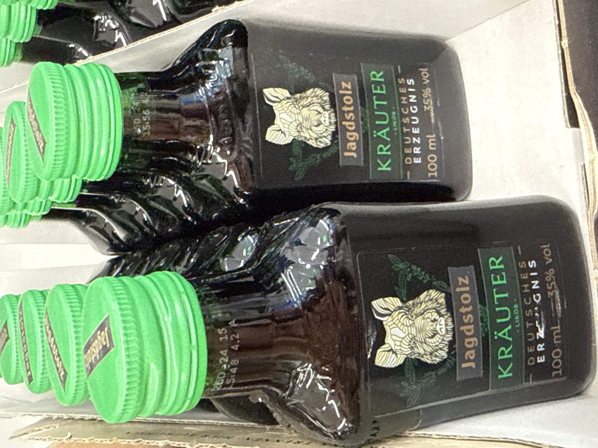 The image shows a close-up of several small, dark glass bottles with bright green caps arranged in a display, likely in a retail setting. Each bottle has a black label featuring a stylized drawing of a wild boar's head, above text that reads "Jagdstolz" and "KRÄUTER". Below this, it states "DEUTSCHES ERZEUGNIS" (German Product), "100 ml", and "35% vol". The bottles are stacked closely together, suggesting they are part of a larger product offering. The lighting is bright, with some glare on the glass bottles. The background is blurred, but appears to be made of cardboard, consistent with product packaging. There are no people or animals visible in the image. The context suggests these are bottles of Jagdstolz Kräuter liqueur, a German herbal liqueur, and given the detail on the label, the location is likely a shop or supermarket in Germany. Without further information, it is impossible to pinpoint the exact location within Herzberg, Germany.
