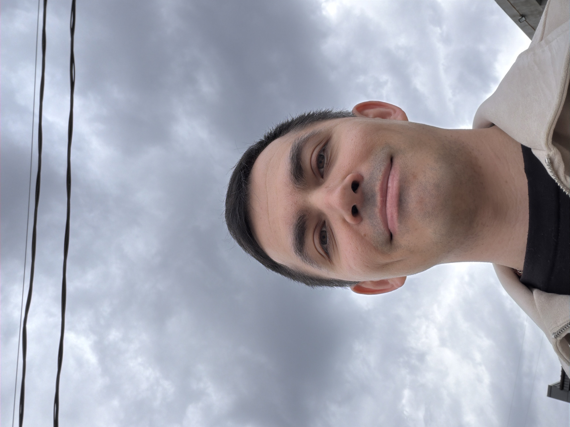 The image shows a medium close-up of a man with dark hair and fair skin. He is wearing a light-colored jacket over a black shirt. The background is a cloudy sky. Electrical wires run along the left side of the frame. The picture is taken at an angle. There is no evidence to determine whether the image was taken in Izhevsk, Russia.