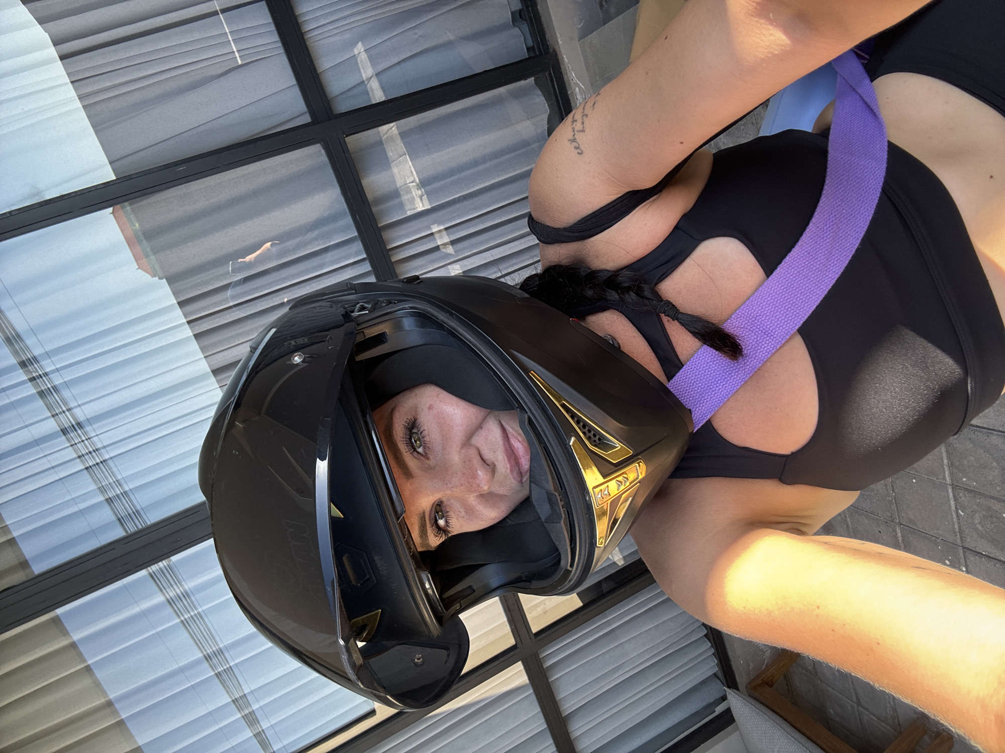 A woman is taking a selfie outdoors, wearing a black motorcycle helmet, black sports bra, and a purple strap across her chest. She has a braid of dark hair and freckles. She is looking directly at the camera. Behind her, large windows with sheer white curtains are visible. The scene appears to be a balcony or patio in North Kuta, Indonesia, suggested by the architectural style and the presence of outdoor furniture. The lighting indicates it is daytime, with strong sunlight casting shadows.