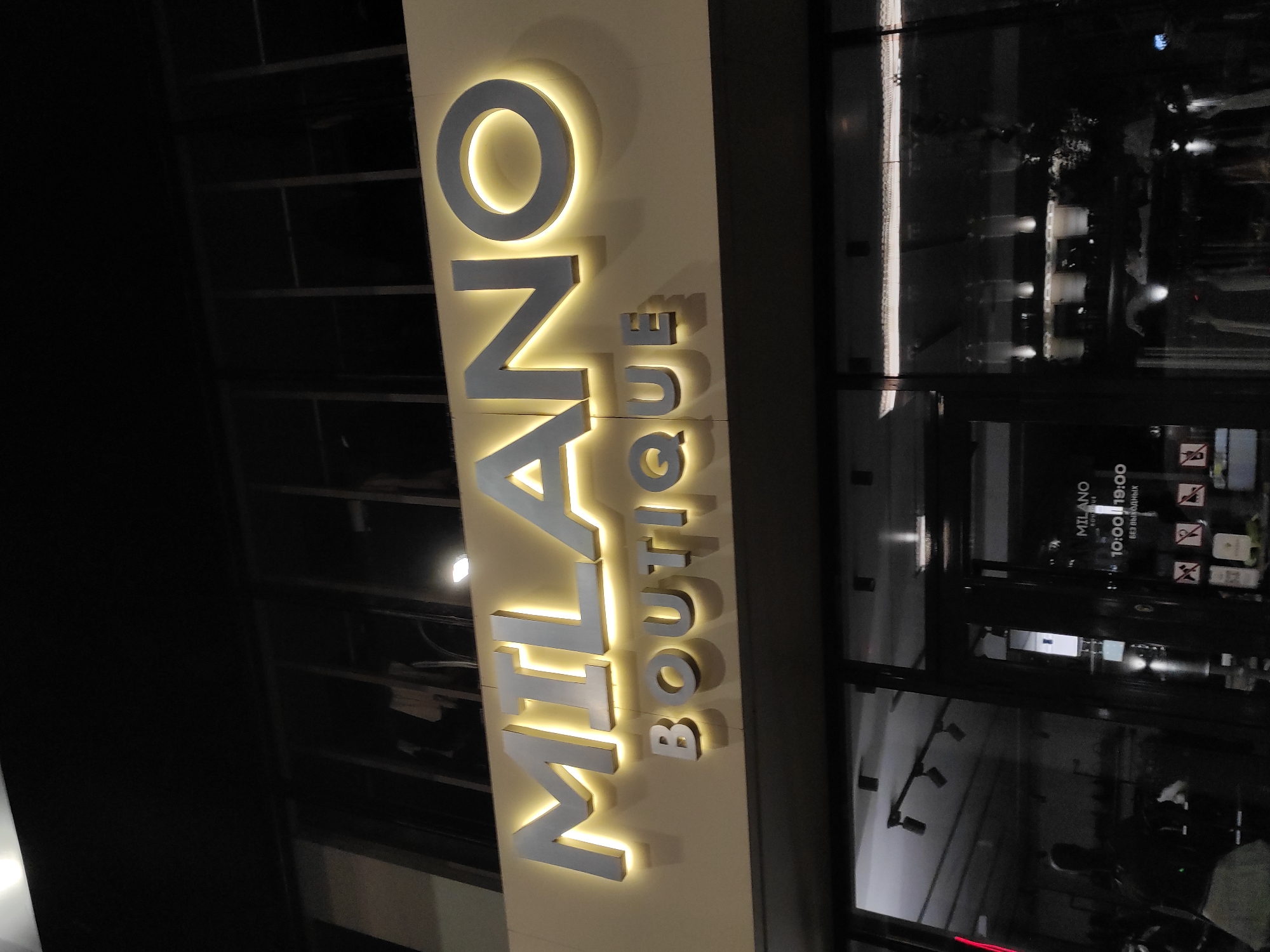 Here is a description of the image:
The image shows a vertical sign for "MILANO BOUTIQUE" with each letter illuminated from behind, located outside a store in an urban environment. The background indicates an outdoor setting at night, with lights and reflections visible through the store's glass windows and door.