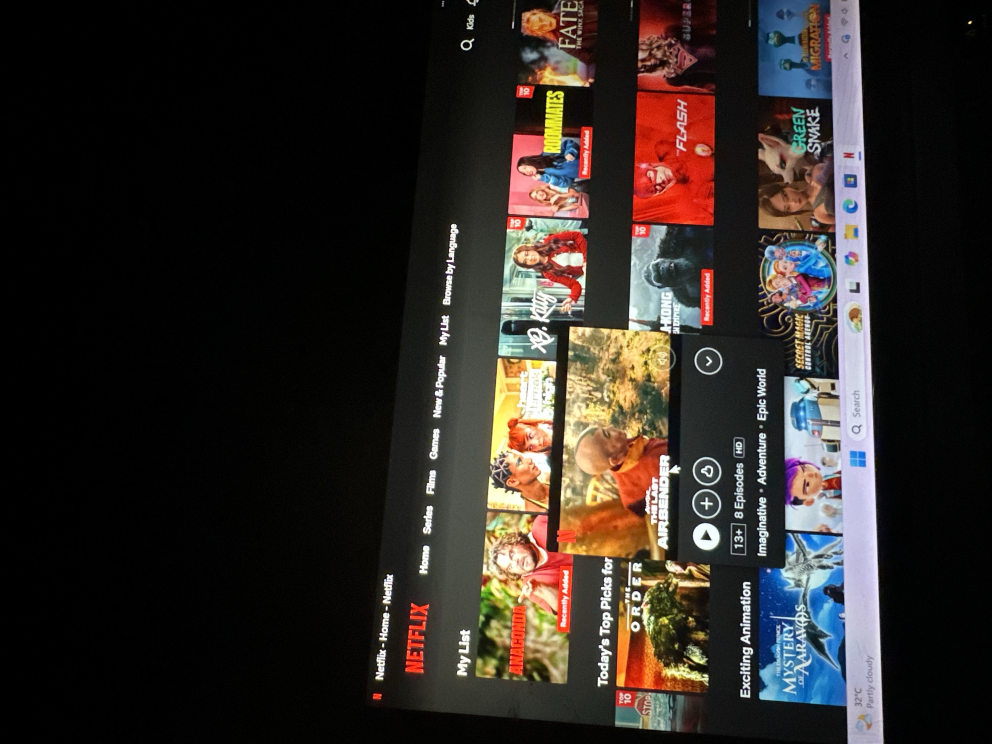 The image captures a digital screen, likely a laptop or monitor, displaying the Netflix streaming service interface. The screen is tilted, with a dark, presumably indoor, background.
The Netflix application occupies the majority of the display, showcasing a grid of movie and TV show thumbnails organized into various categories. Visible navigation options on the left include "Home," "Series," "Films," "Games," "New & Popular," "My List," and "Browse by Language." A search icon and a "Kids" profile option are also present at the top.
Numerous titles of films and series are visible on the screen. These include "FATE THE WINX SAGA," "ROOMMATES," "Xo, Kitty," "FLASH," "A-KONG EN EMPIRE" (likely Kong: Skull Island), "ANACONDA," "THE ORDER," "AVATAR THE LAST AIRBENDER," "SECRET MAGIC CONTROL AGENCY," "GREEN SNAKE," "MIGRATION," and "THE DRAGON PRINCE MYSTERY OF AARAVOS." Some titles are highlighted with "Recently Added" or "TOP 10" labels. For "AVATAR THE LAST AIRBENDER," additional details like "13+ 8 Episodes HD" and genre tags "Imaginative Adventure Epic World" are displayed. A cursor, depicted as a white hand icon, is visible over one of the thumbnails.
A segment of the Windows taskbar is visible at the bottom right, displaying various application icons and current weather information: "32°C Partly cloudy." This warm temperature aligns with the typical climate of Kirana, Nigeria.