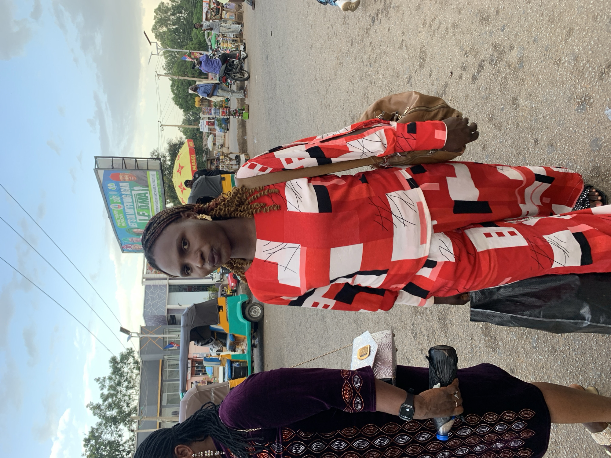 The image is a medium shot of two women standing on a street in Jos, Nigeria. The woman on the right is wearing a red and white patterned jumpsuit and a brown shoulder bag. She is looking at the camera with her mouth slightly open. The woman on the left is wearing a dark purple dress with embellishments and carrying a silver purse and a black plastic bag. She is holding a bottle of water in her right hand. In the background, there are several auto-rickshaws, motorcycles, and people walking. A large billboard with a political advertisement is visible in the upper right quadrant of the image. The sky is cloudy, suggesting it might be late afternoon or early evening. The overall scene depicts a busy street in a Nigerian city, likely a market or transport hub.